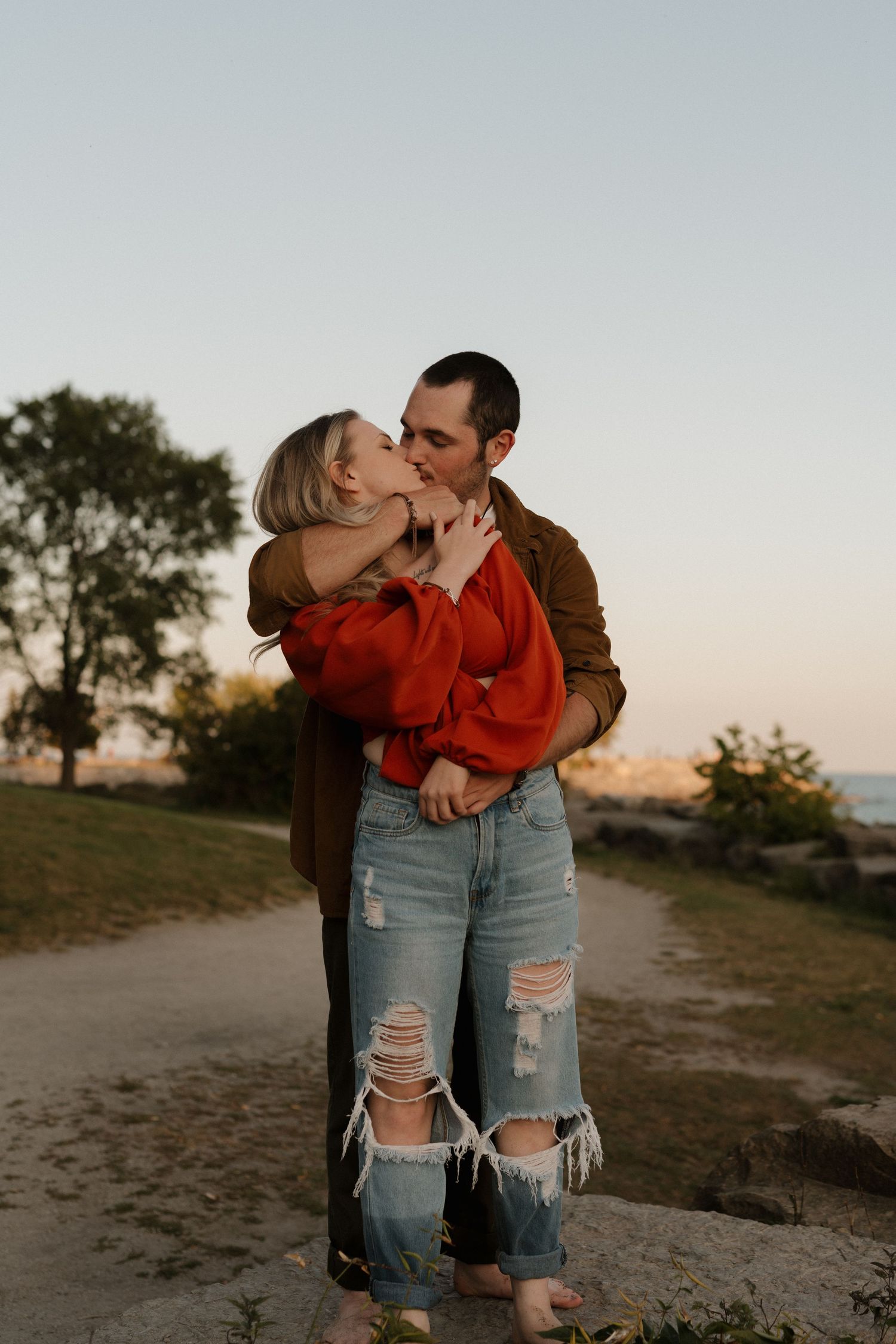A romantic couple sharing a tender kiss at sunset on a dirt path with trees in the background.