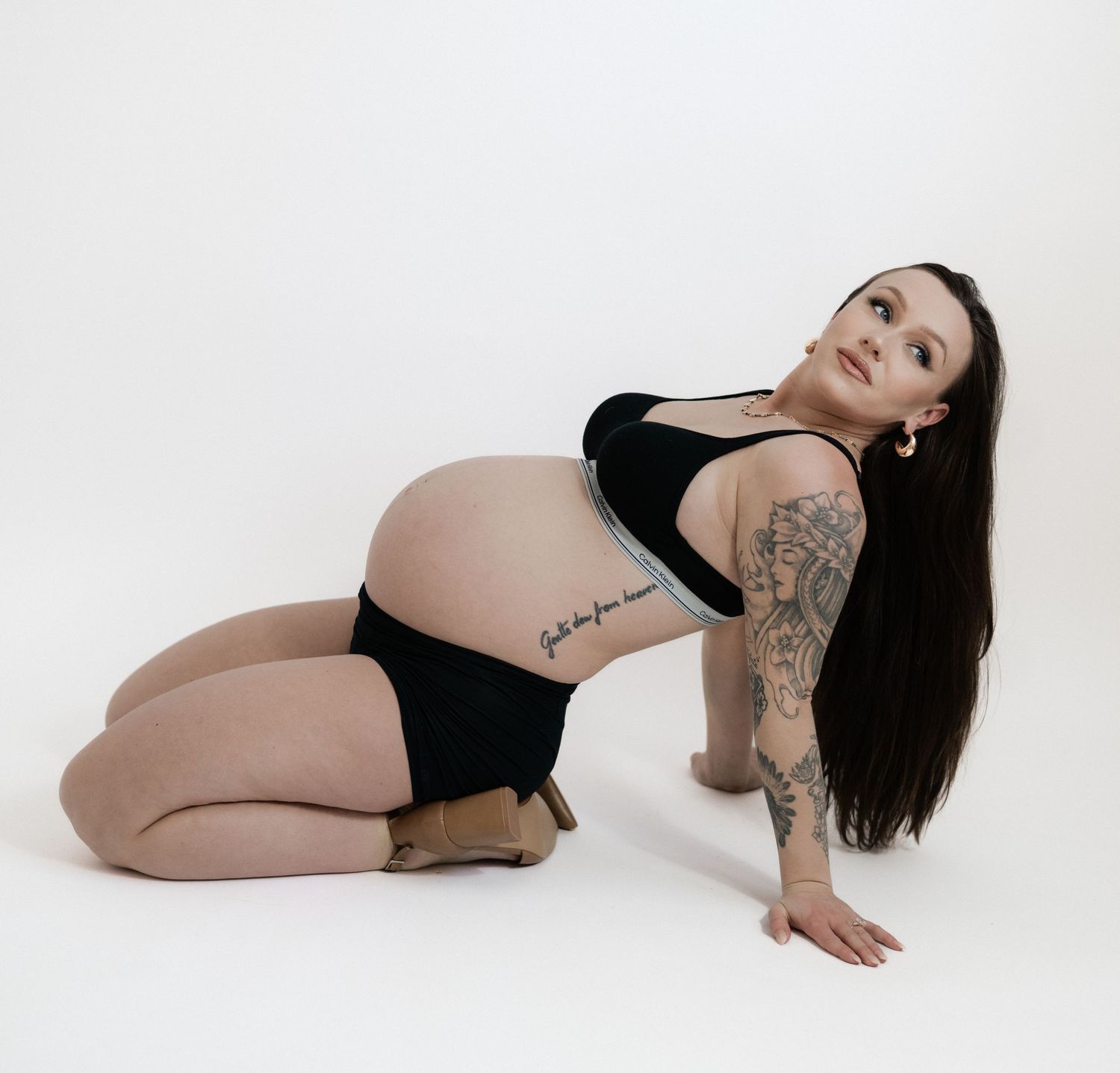 Pregnant woman with tattoos posing in black swimwear on white background, showcasing baby bump.