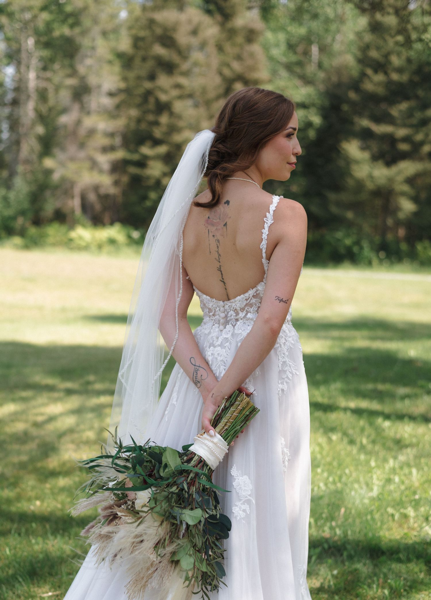 A bride in a detailed white gown poses with her bouquet in a meadow with evergreen trees.