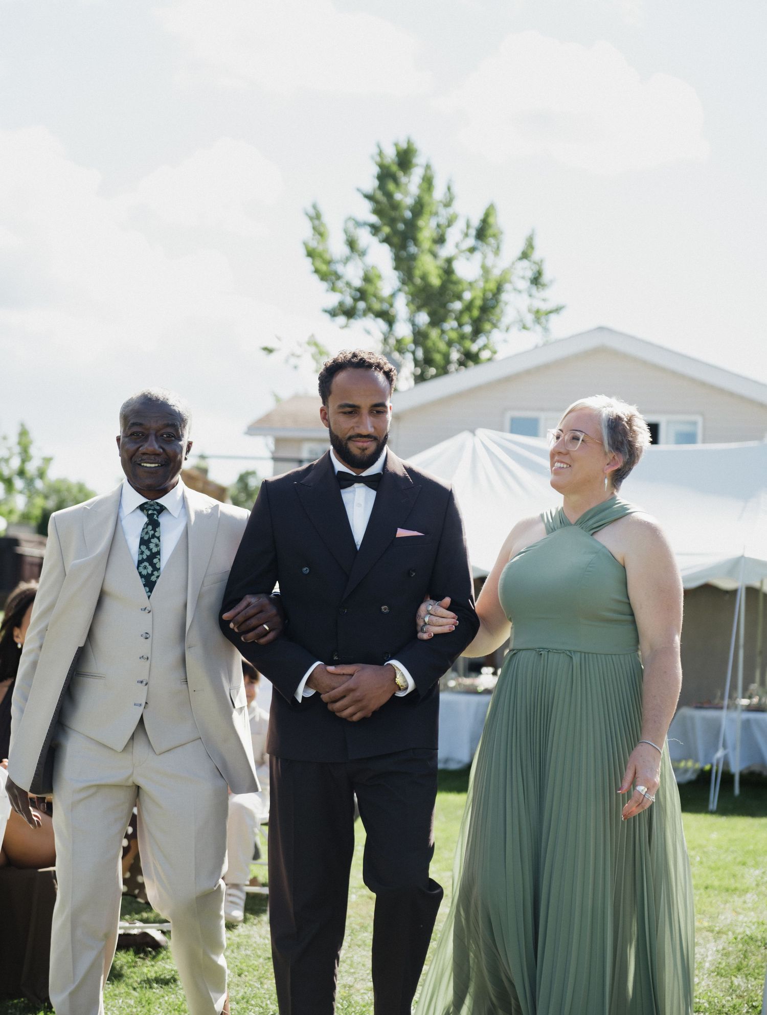 Groom in black tuxedo walks with parents at outdoor wedding reception on sunny day.
