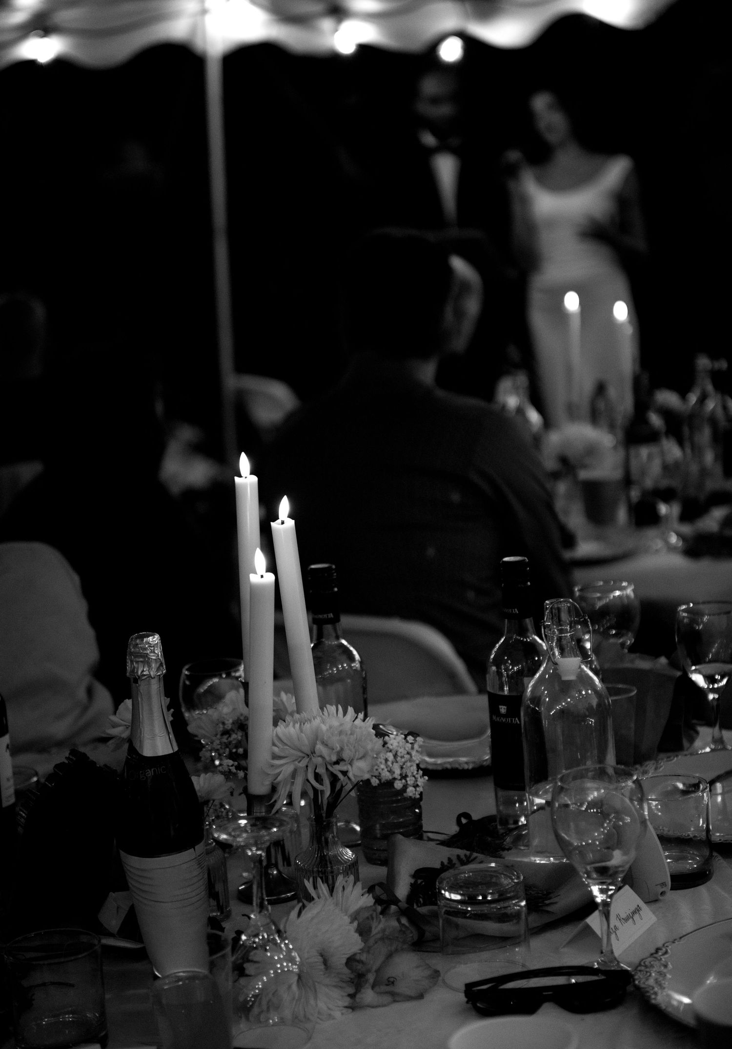 Black and white photo of an elegant candlelit wedding reception table with champagne, flowers, and guests in formal attire.