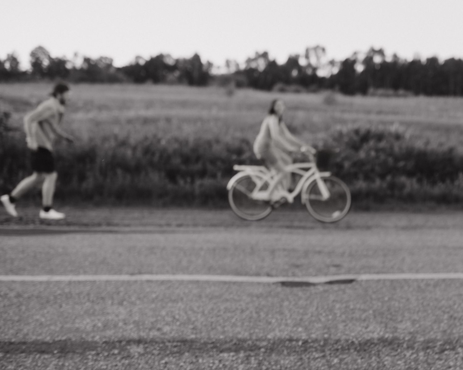 Black and white photo of a woman riding a bicycle and a man jogging along a rural road with fields in the background.