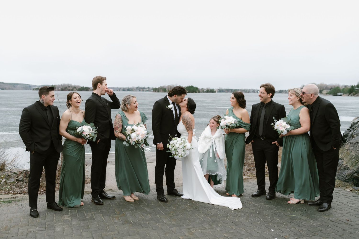 Wedding party poses by waterfront, bridesmaids in green dresses, groomsmen in black, as bride and groom share a kiss.