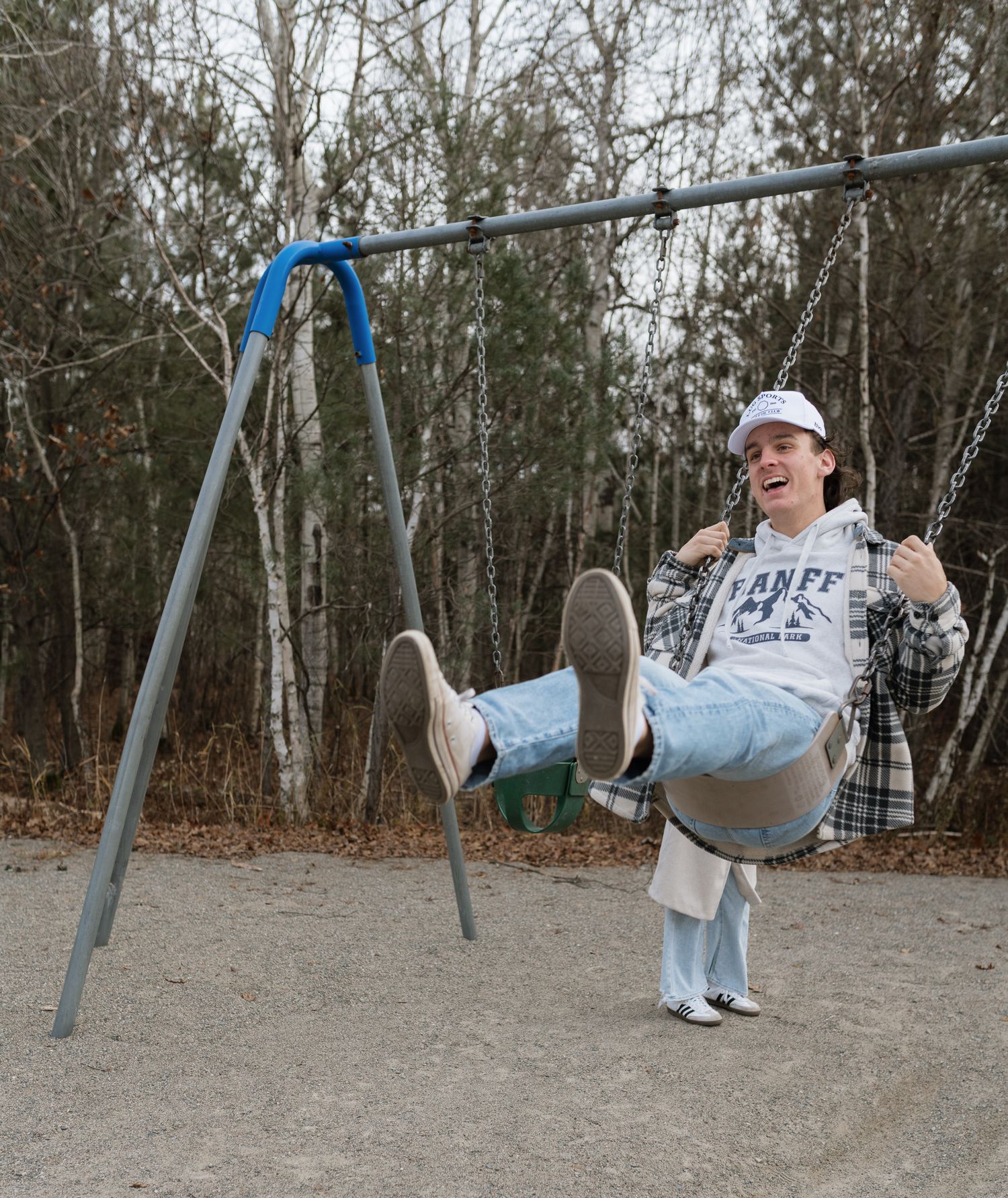 A laughing person swinging on a playground swing set outdoors in autumn, wearing a cap and casual clothes.