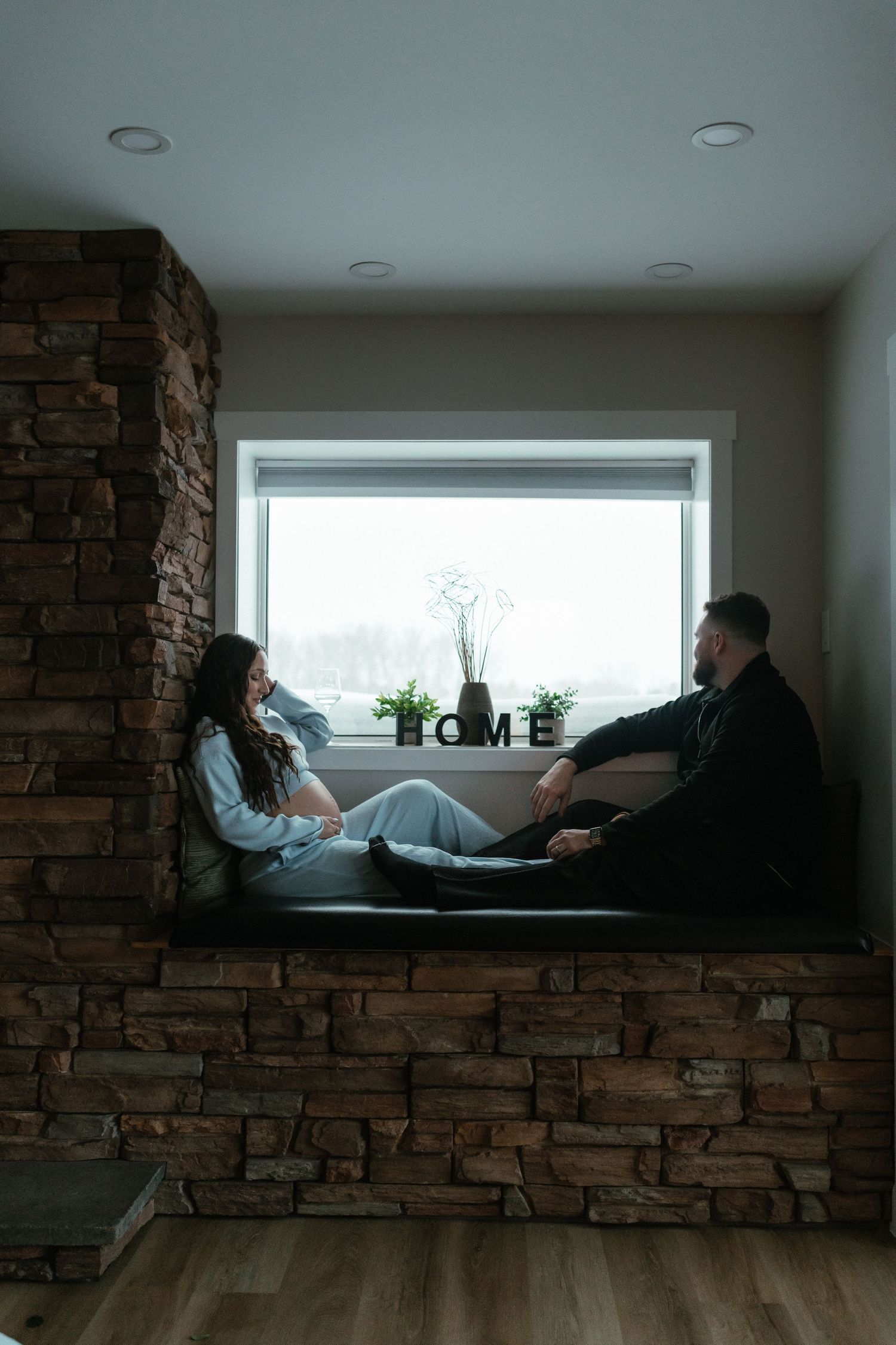 Couple relaxing on a stone window seat with HOME letters and snowy view outside.