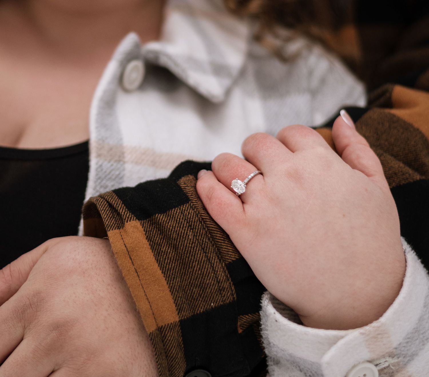 Close-up of a woman's hand wearing a diamond solitaire engagement ring, resting on a plaid flannel shirt.