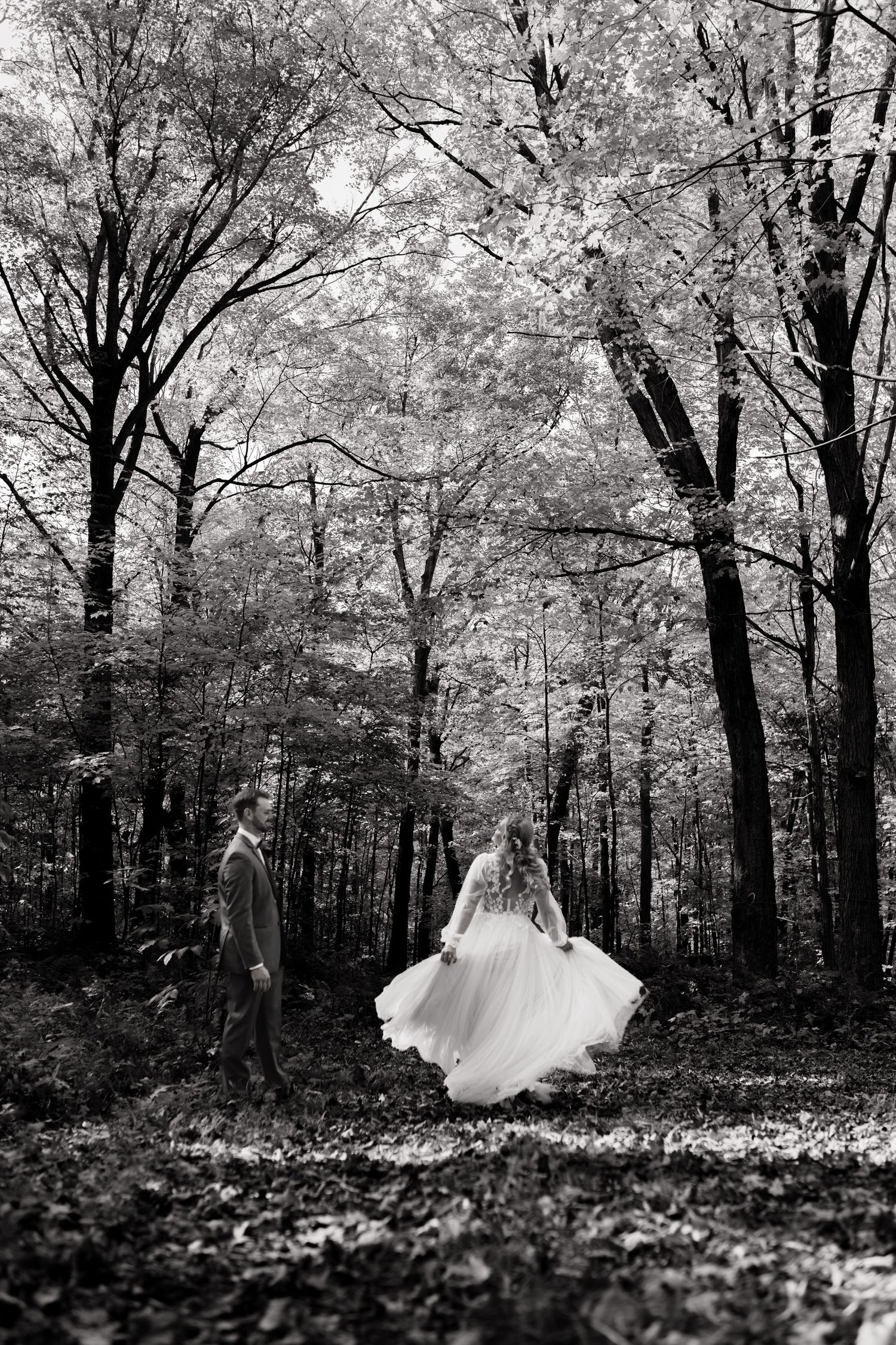 Black and white photo of a bride in flowing gown and groom walking through a lush forest during their wedding day.