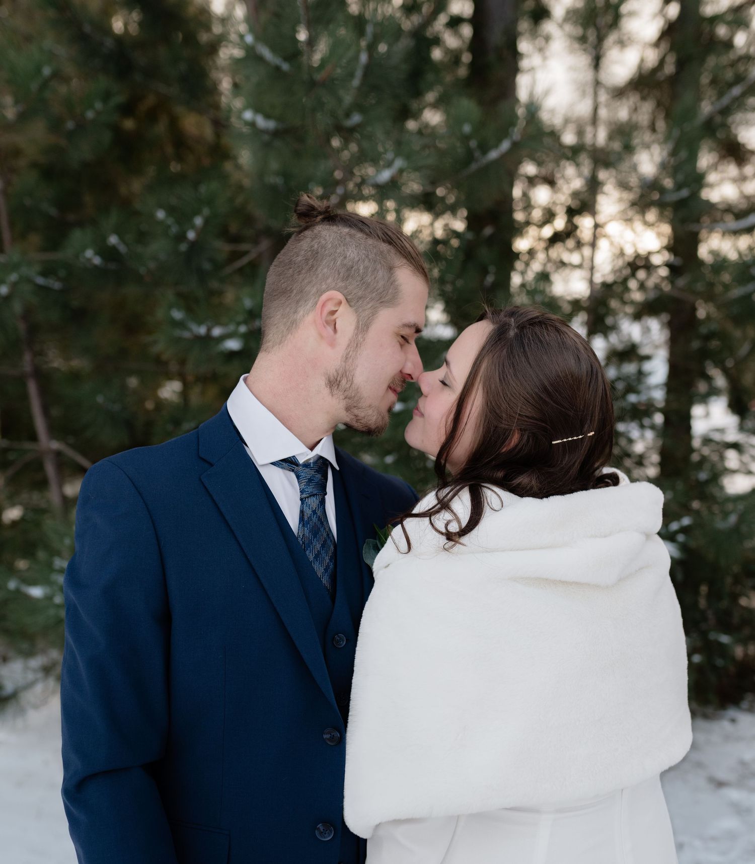 A winter wedding moment showing a couple in navy suit and white wrap embracing in a snowy forest.