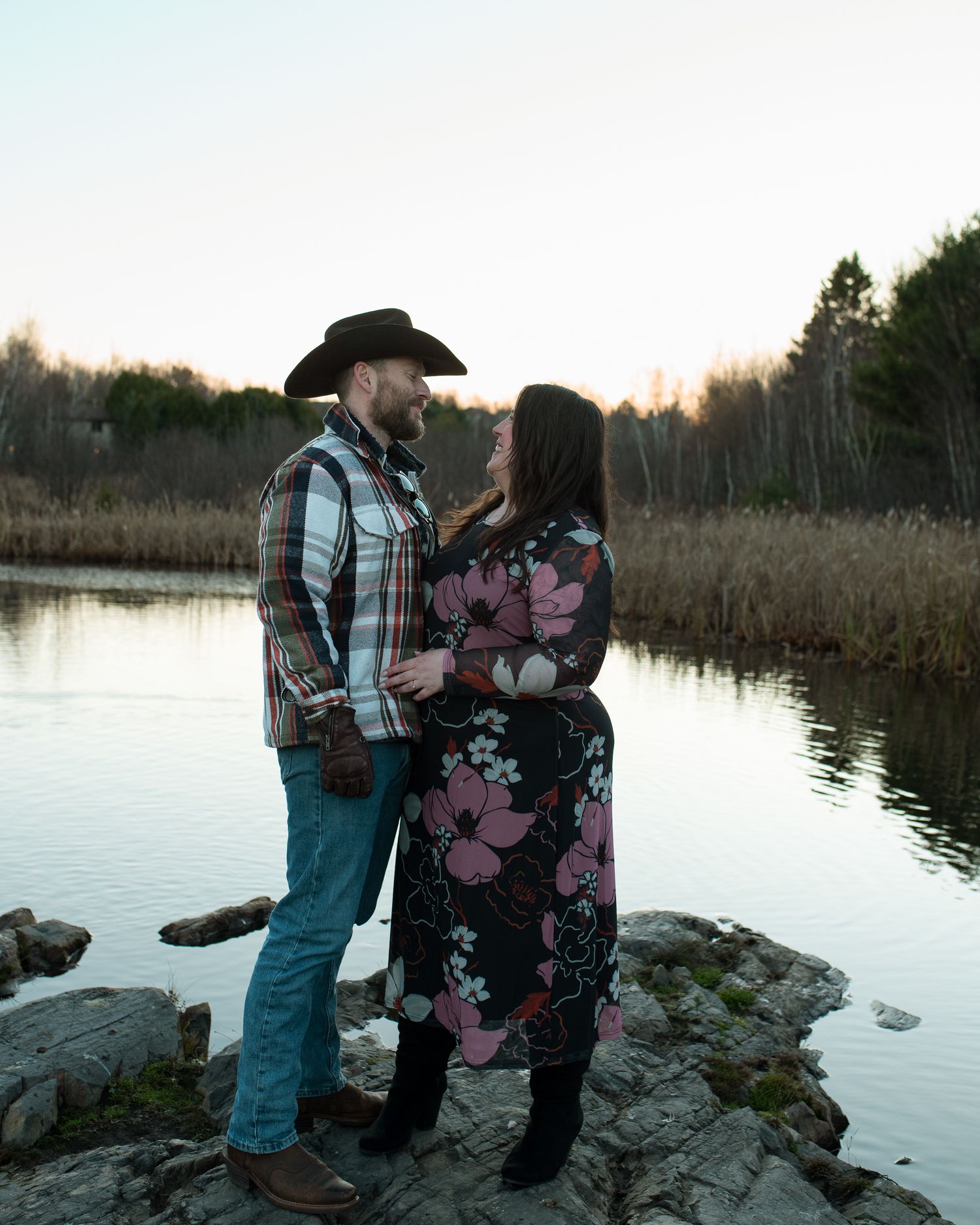 A couple shares a tender moment by a serene lake at sunset while standing on rocky shoreline in winter.