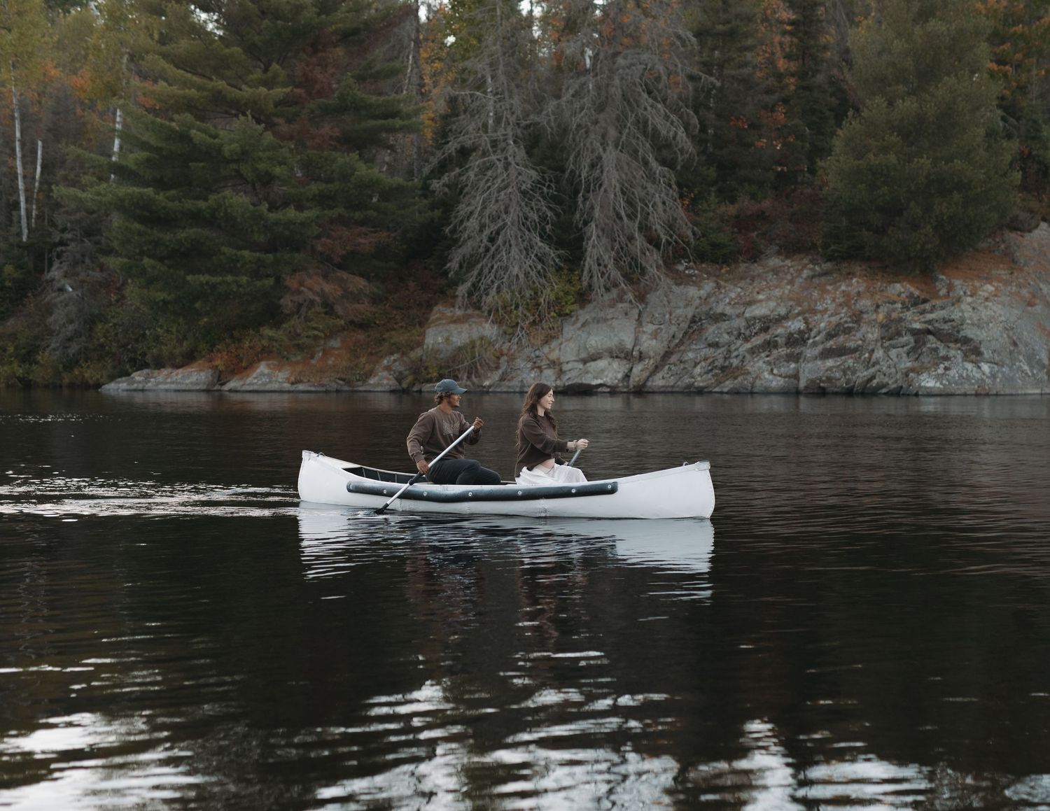 A white canoe glides across a serene lake surrounded by autumn trees and rocky shoreline.