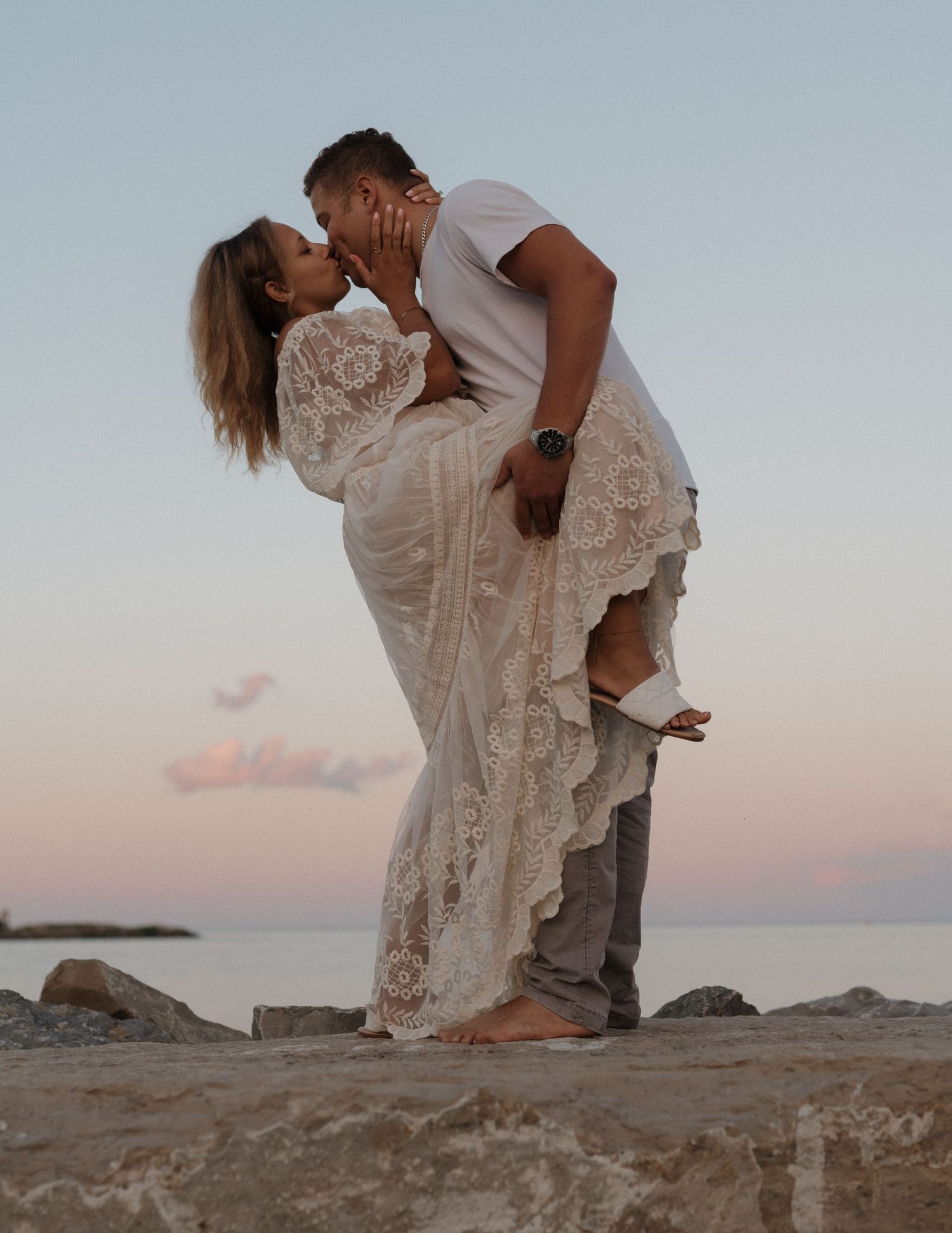 A romantic couple embraces on a rocky beach during sunset, with her wearing a flowing lace dress.