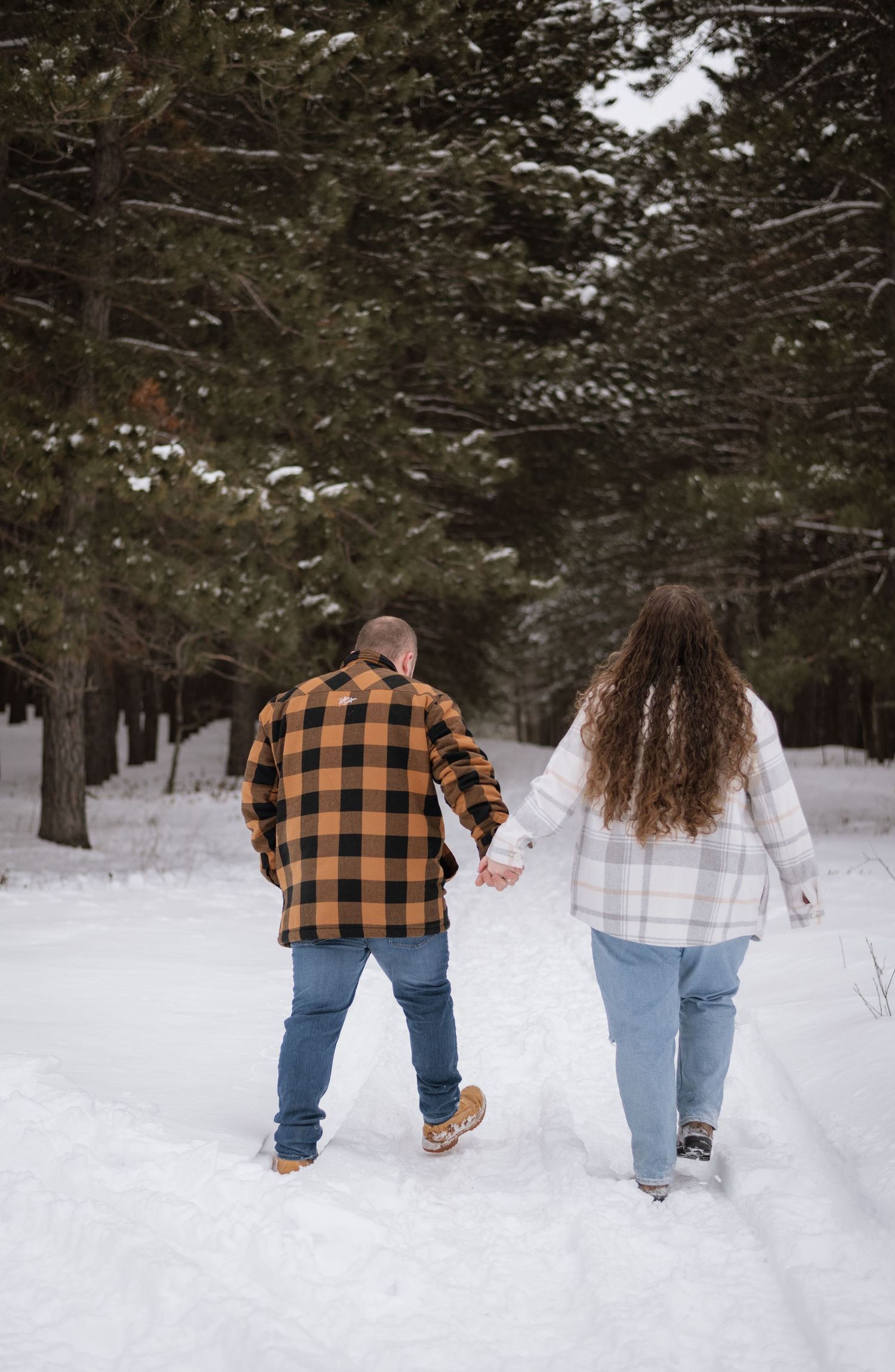 Couple holding hands walking through a snowy forest path in plaid shirts and jeans during winter.