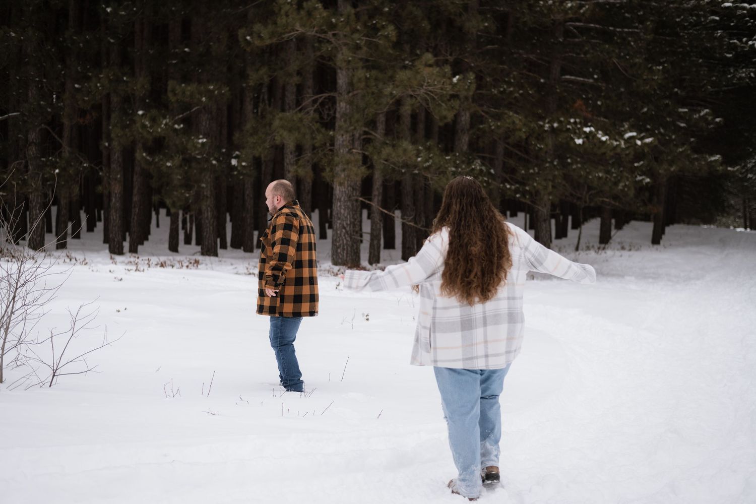 Couple walking in snowy winter forest clearing, wearing flannel shirts and jeans among tall pine trees.