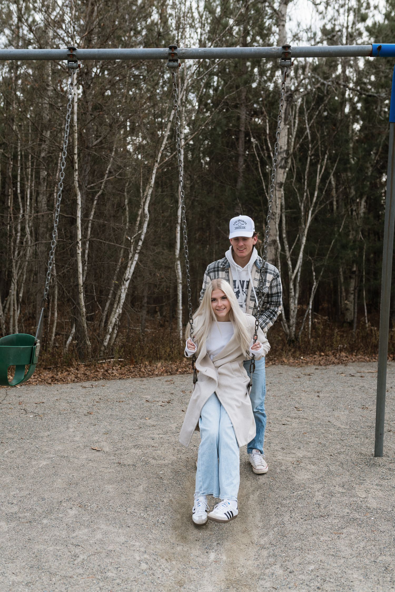 Young couple at playground, man pushing blonde woman on swing, wearing casual fall outfits.