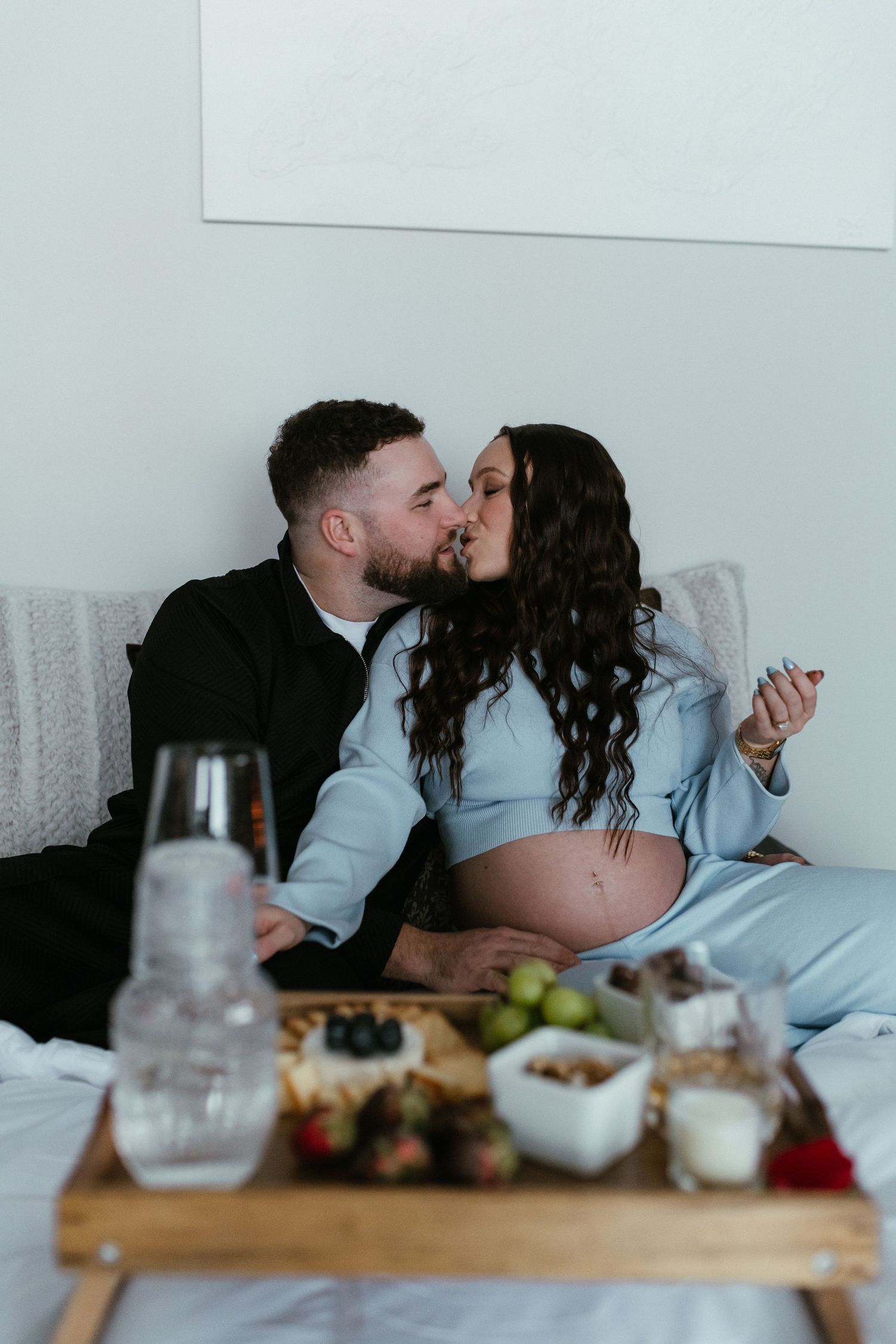 Pregnant couple sharing a kiss in bed with a wooden breakfast tray of fruits and snacks in the foreground.