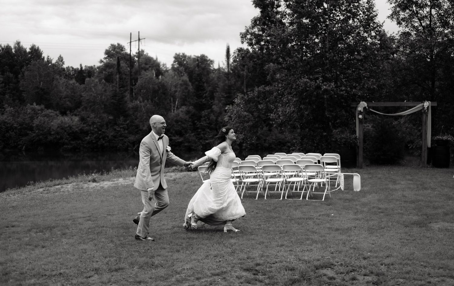 Black and white photo of a bride and groom dancing joyfully on a grass lawn with white chairs and trees in the background.