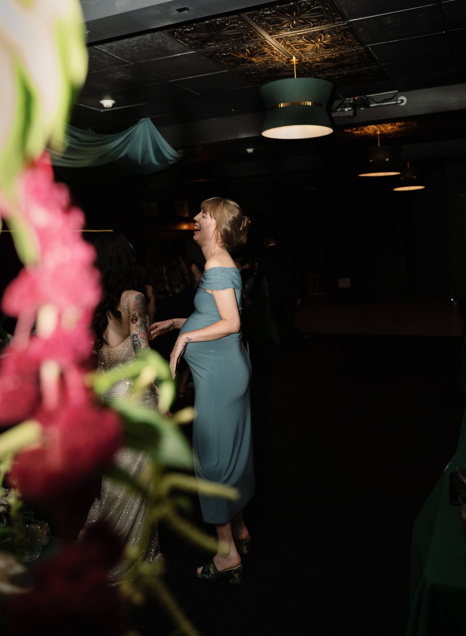 Woman in teal off-shoulder midi dress standing at an elegant dark reception venue with floral arrangements.