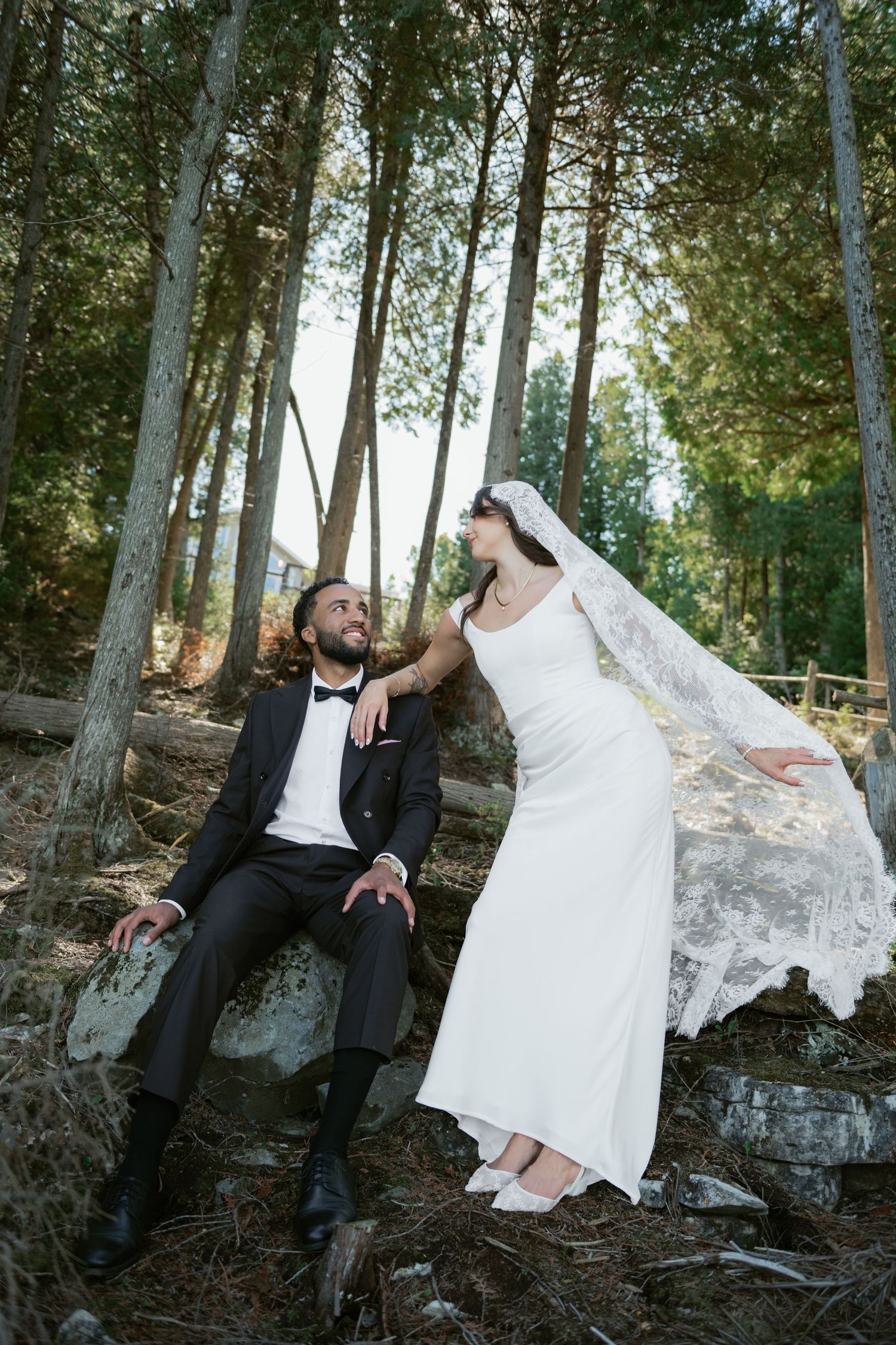 Bride and groom pose on rocks in forest, bride's veil flowing in the wind.