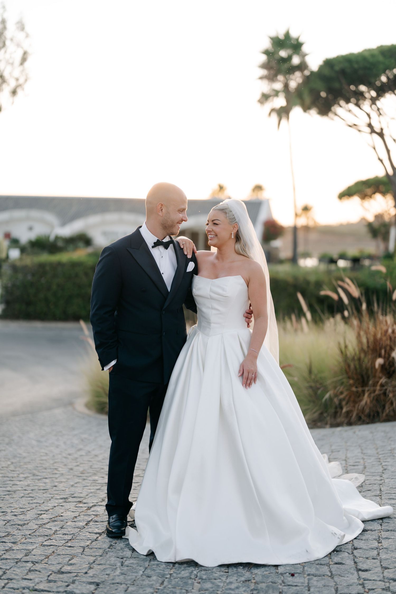 A romantic wedding couple poses together on a stone pathway during golden hour sunset.