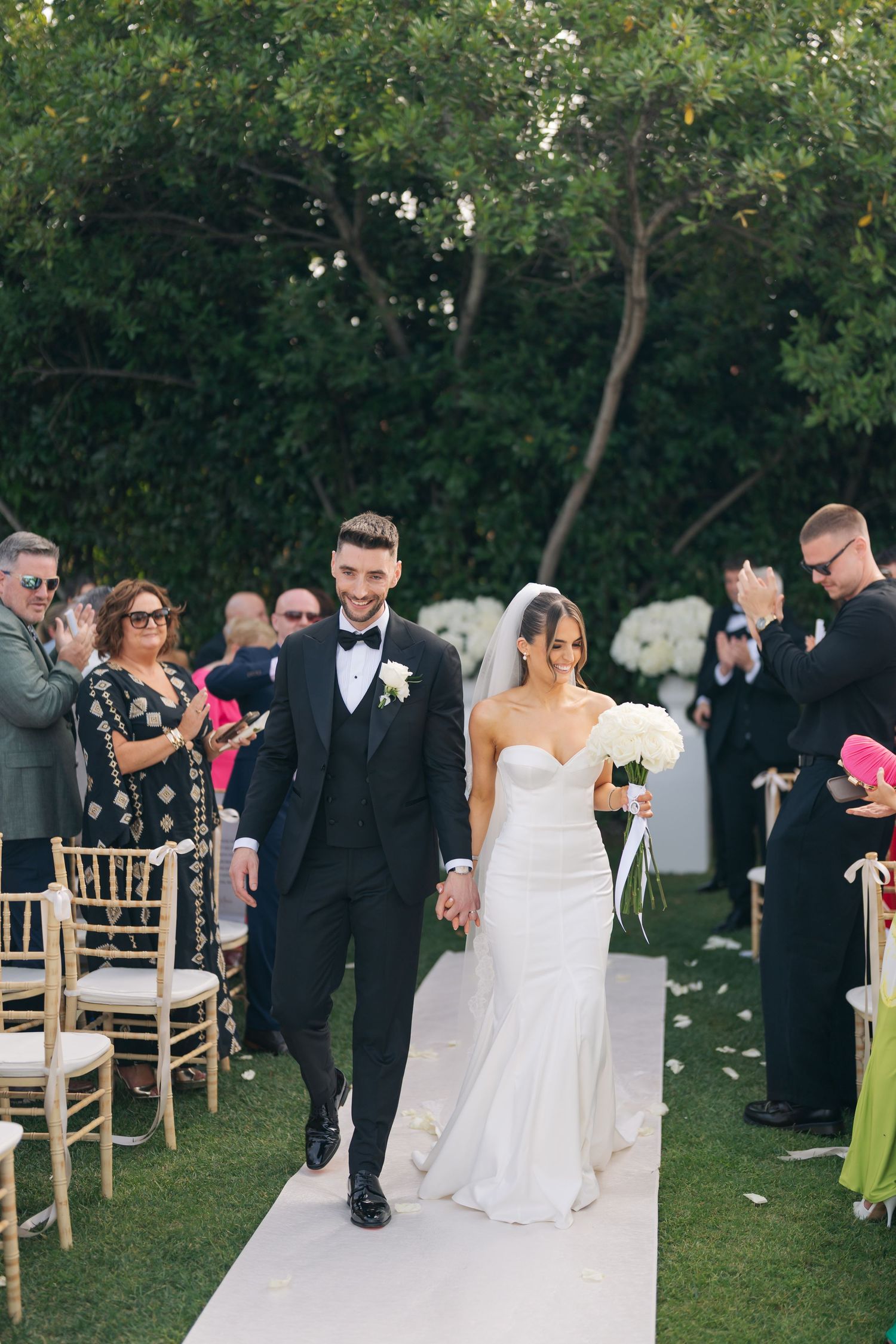 Wedding ceremony processional on white aisle runner with guests seated on gold chairs.