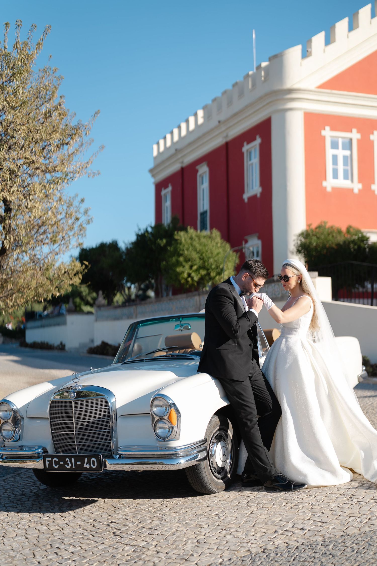 Wedding couple poses with vintage Mercedes-Benz against coral-colored castle architecture.