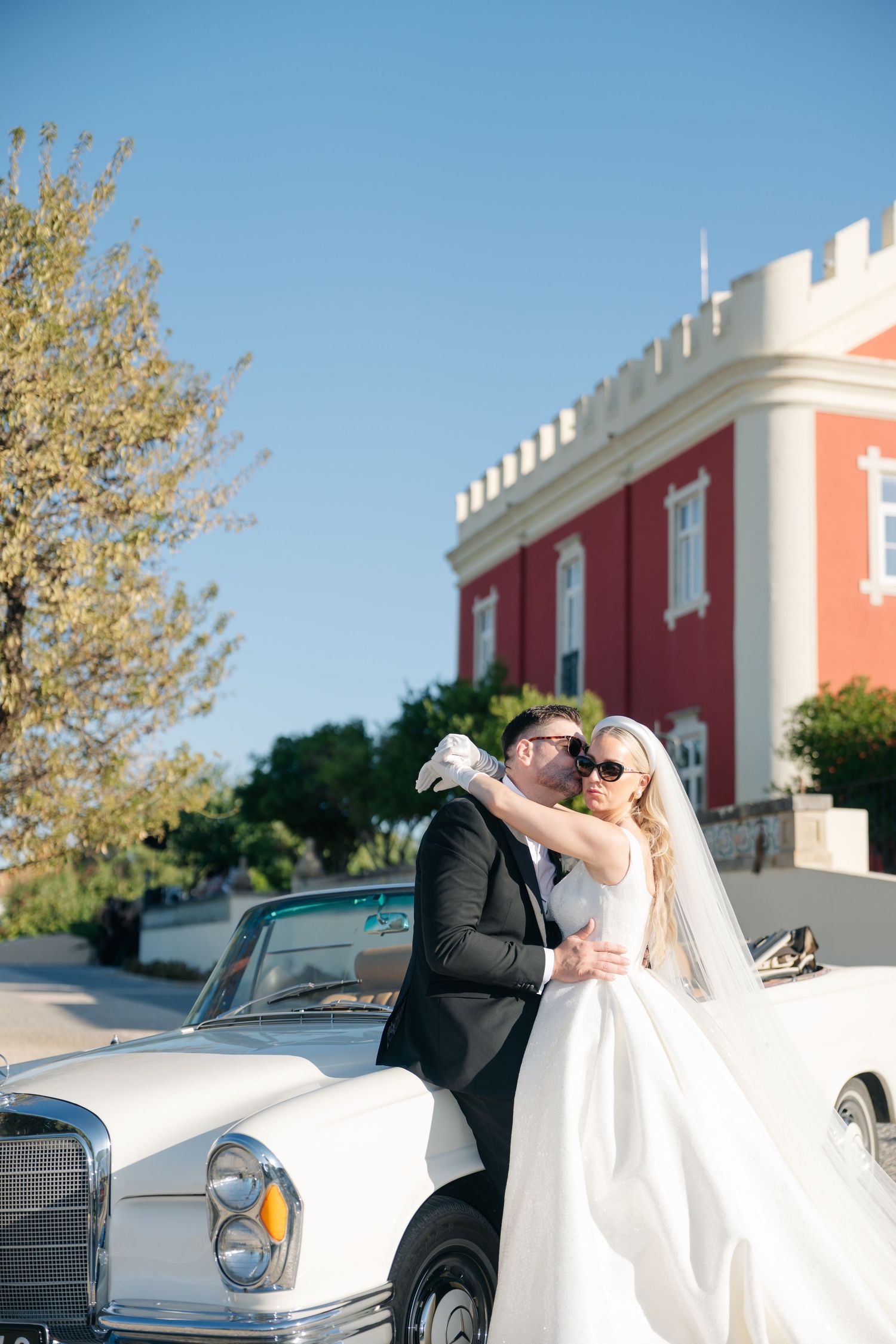 Couple poses near vintage car outside coral-colored building during wedding photoshoot.