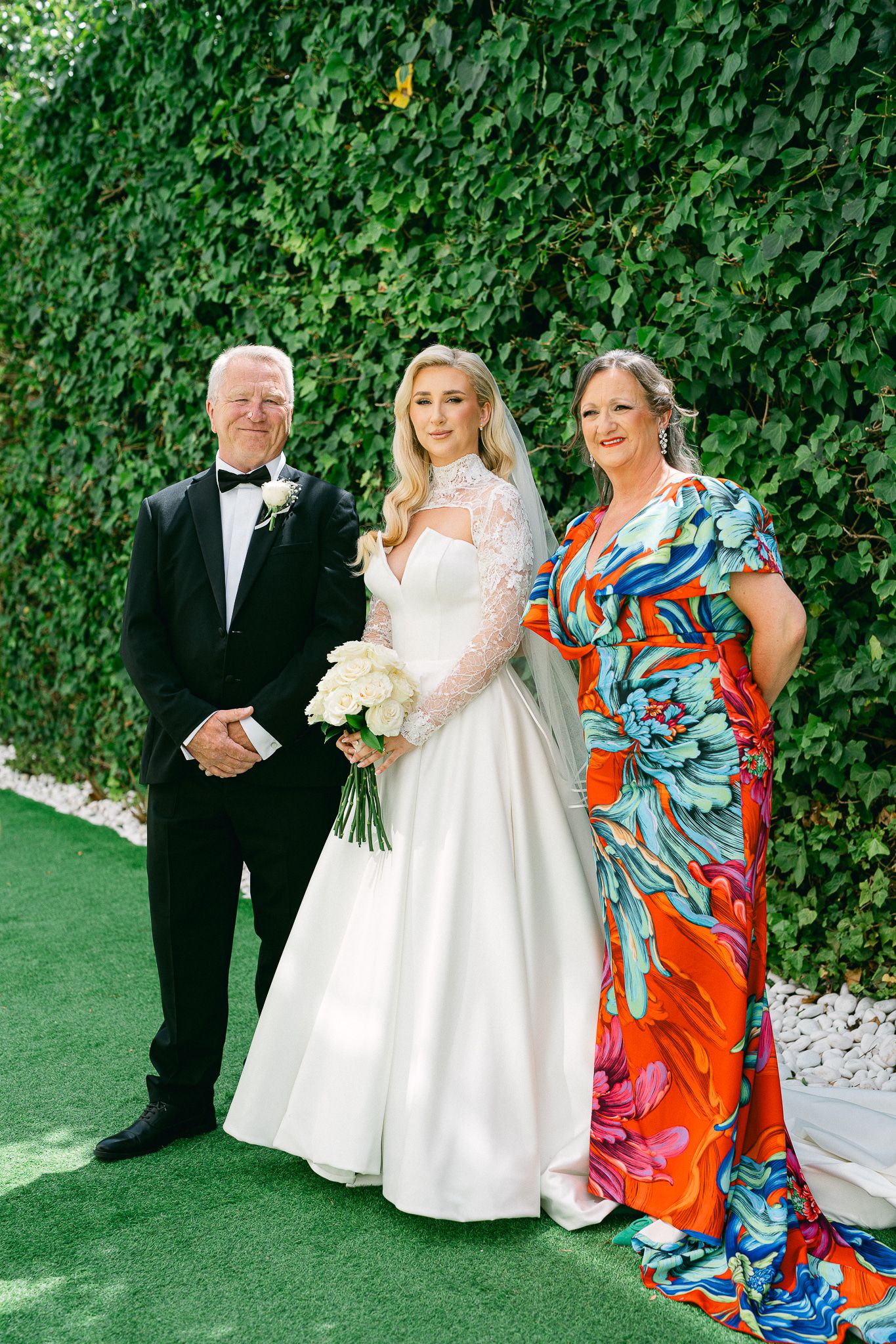 Wedding party poses against an ivy wall with bride in white and guests in colorful attire.