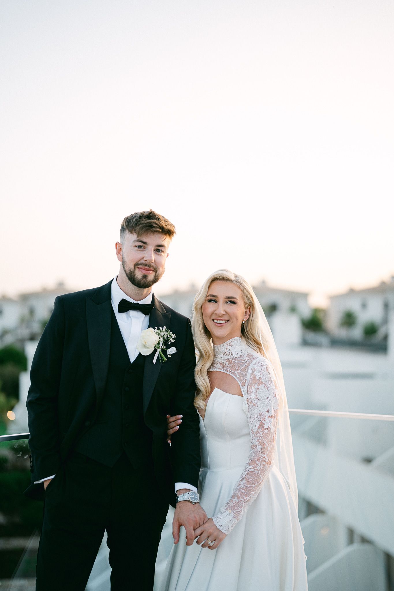 Elegant couple in wedding attire poses at sunset against white building backdrop.