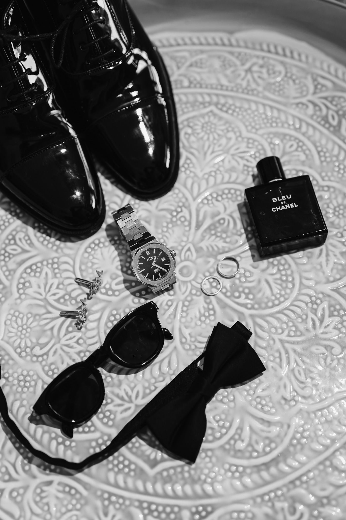 Black dress shoes and accessories arranged on a decorative patterned surface.