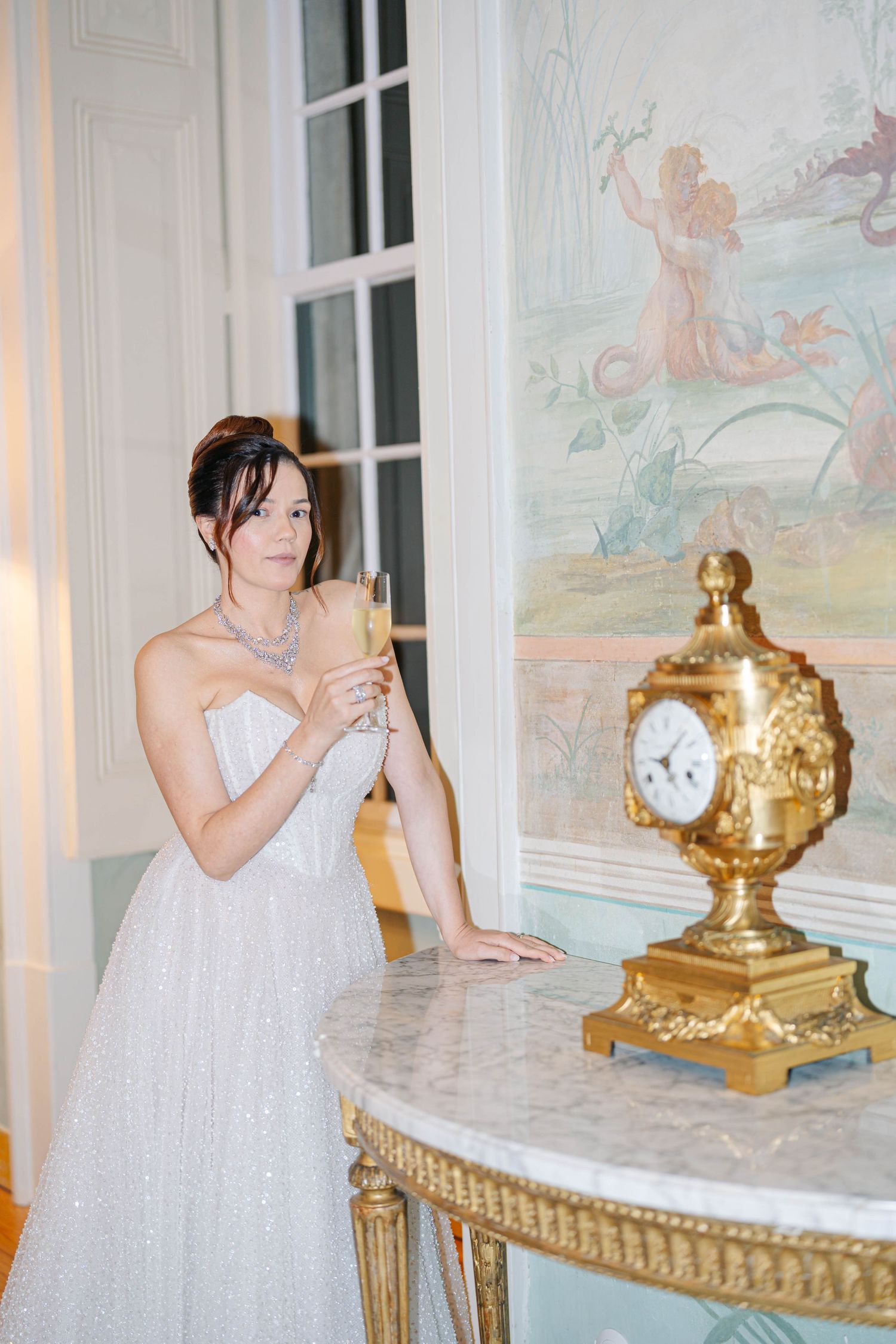A person in a white dress stands by an ornate marble table with gold accents and decorative clocks in an elegant room.