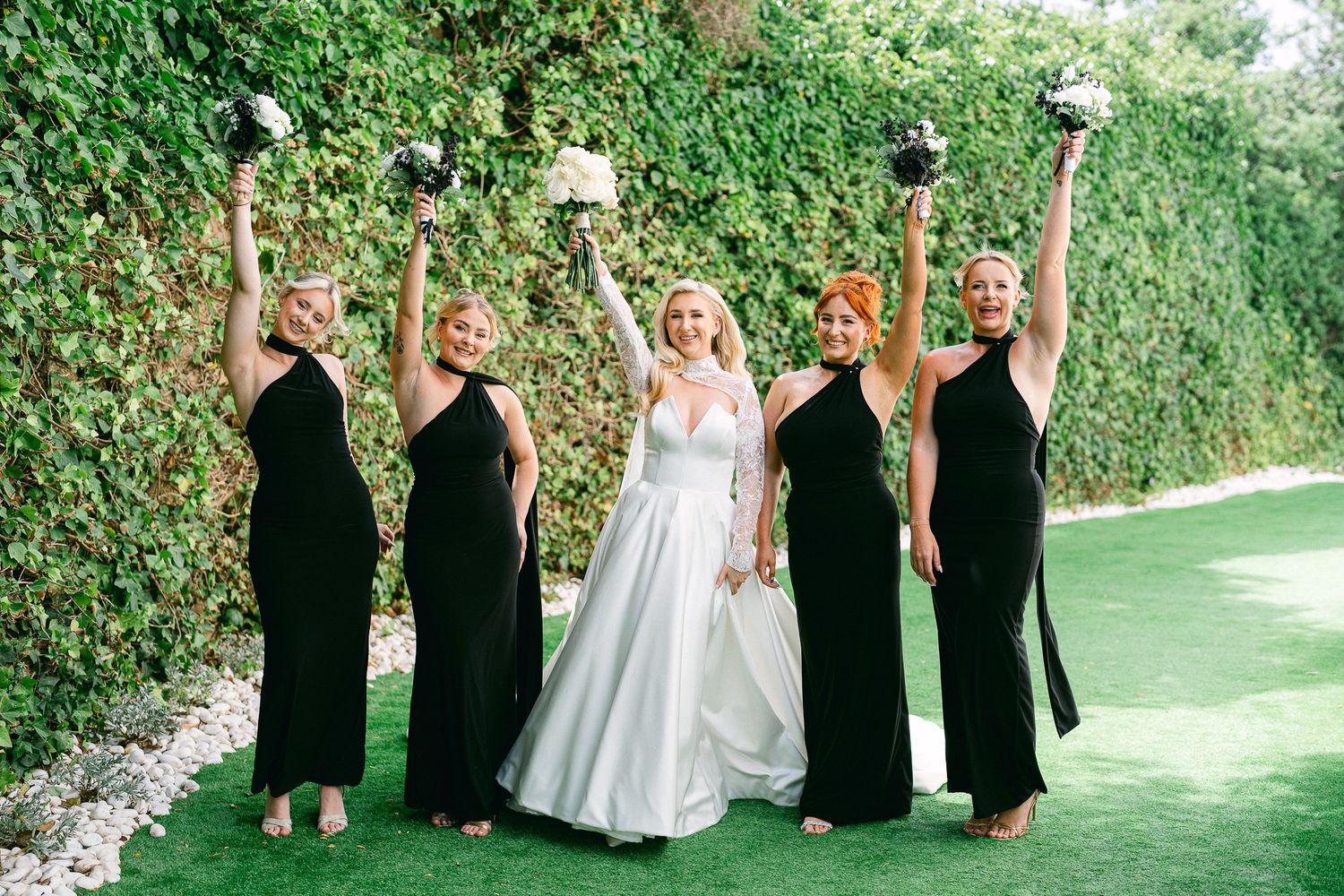 Wedding party members in black and white attire stand in line against green garden backdrop.