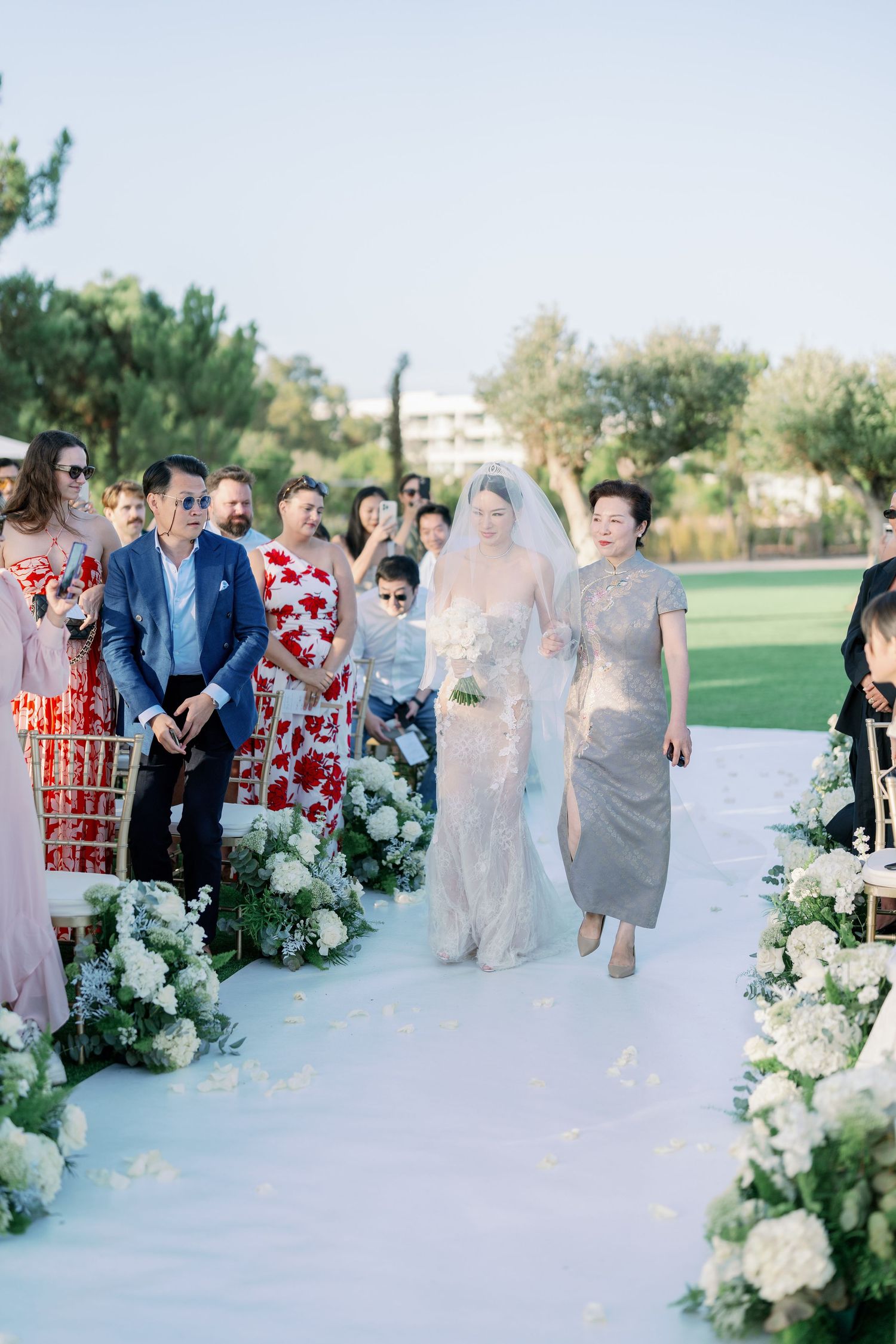 Wedding ceremony aisle decorated with white floral arrangements and guests seated outdoors.