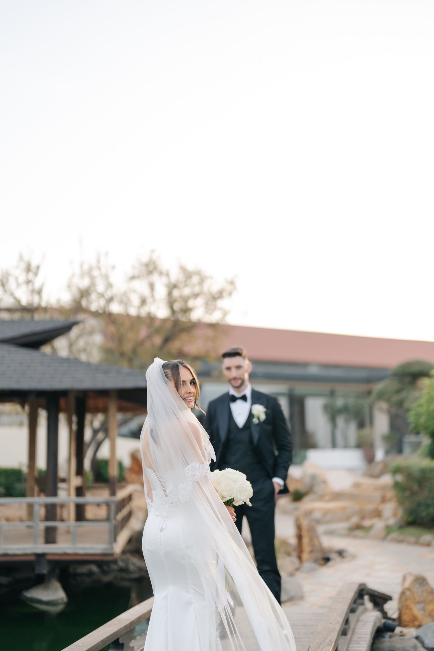 Bride in white gown poses on wooden walkway with flowing veil in afternoon light.
