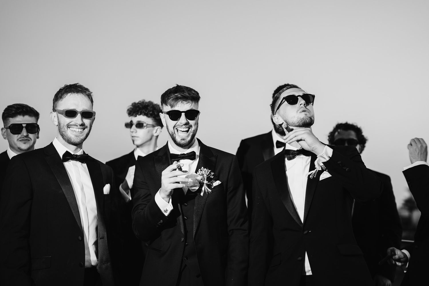 Wedding party members in black suits and sunglasses pose together outdoors.