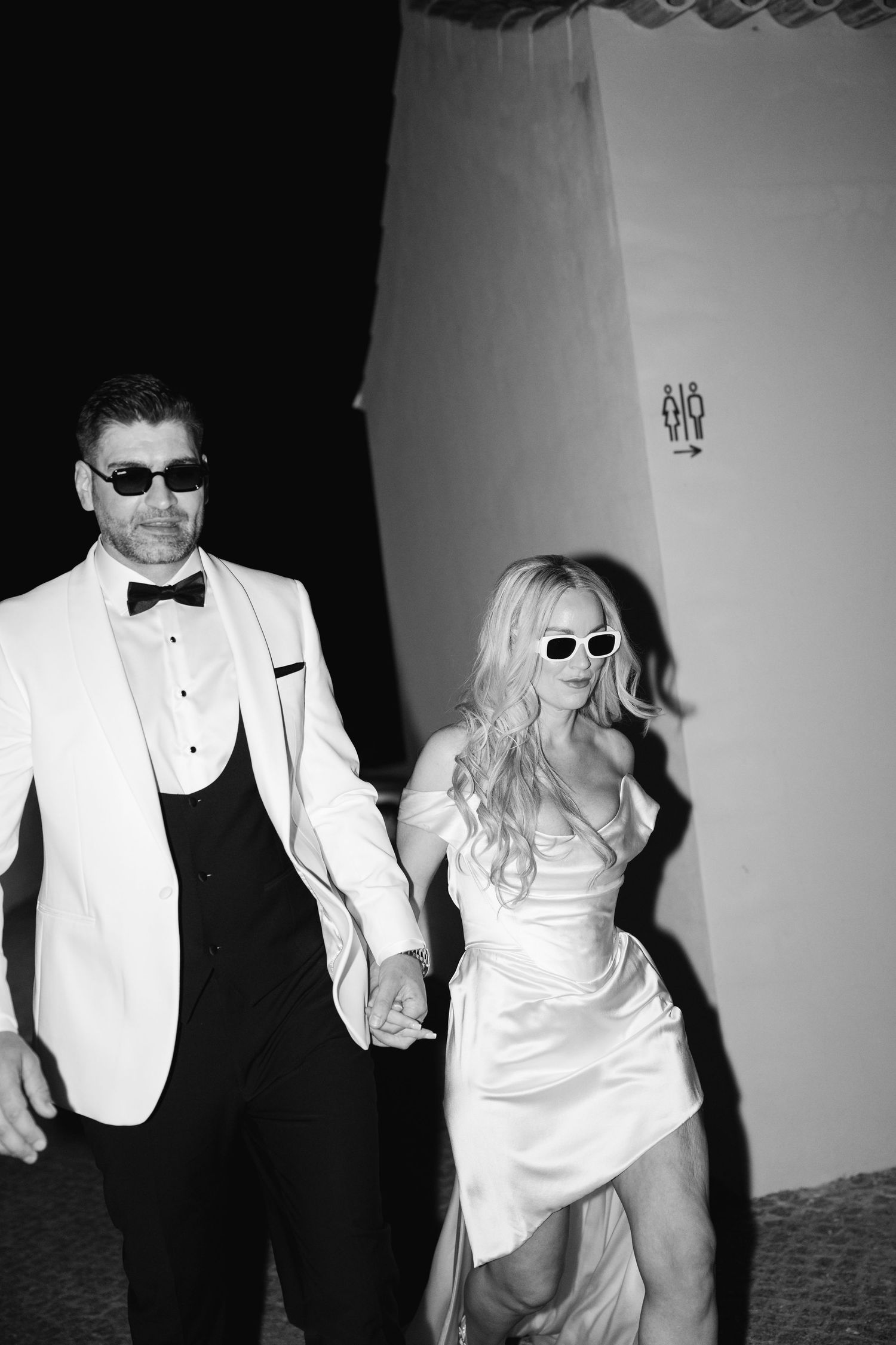 A couple in formal wear walking through a hallway wearing sunglasses.