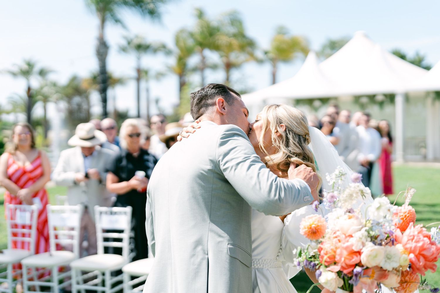 Wedding celebration with guests mingling outdoors under white tents and palm trees.