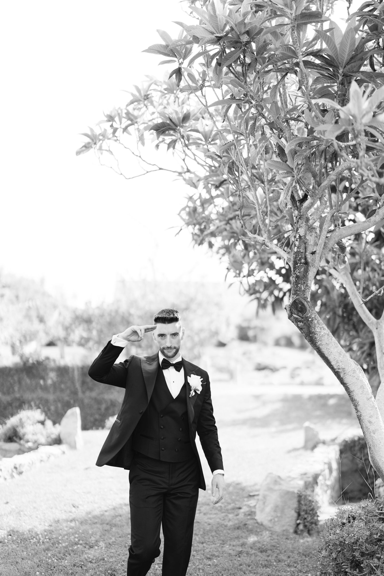 A groom in a black tuxedo walks through a garden setting in black and white photography.