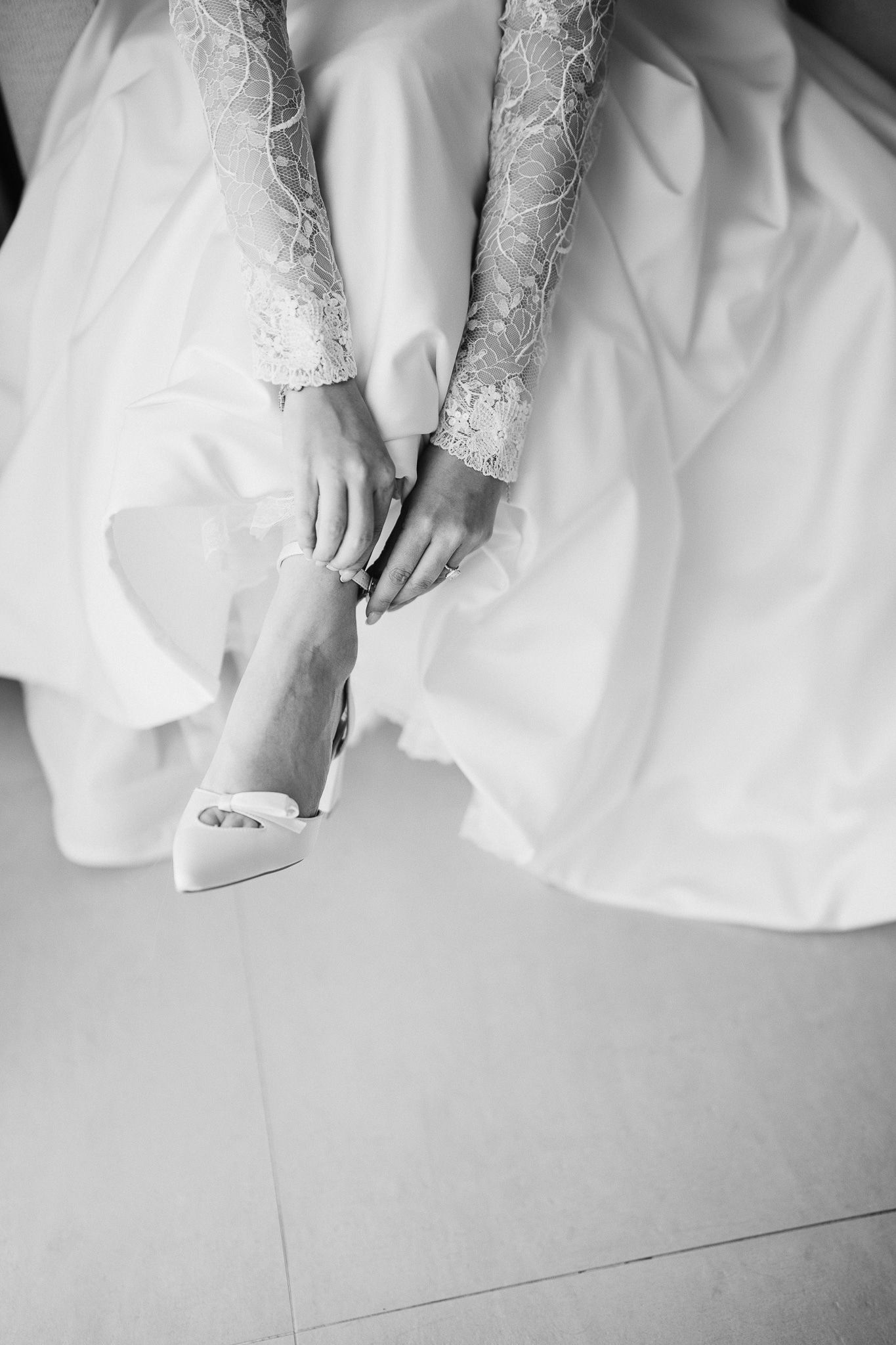 Black and white detail shot of bride adjusting her shoe, showing delicate lace sleeves and full skirt.
