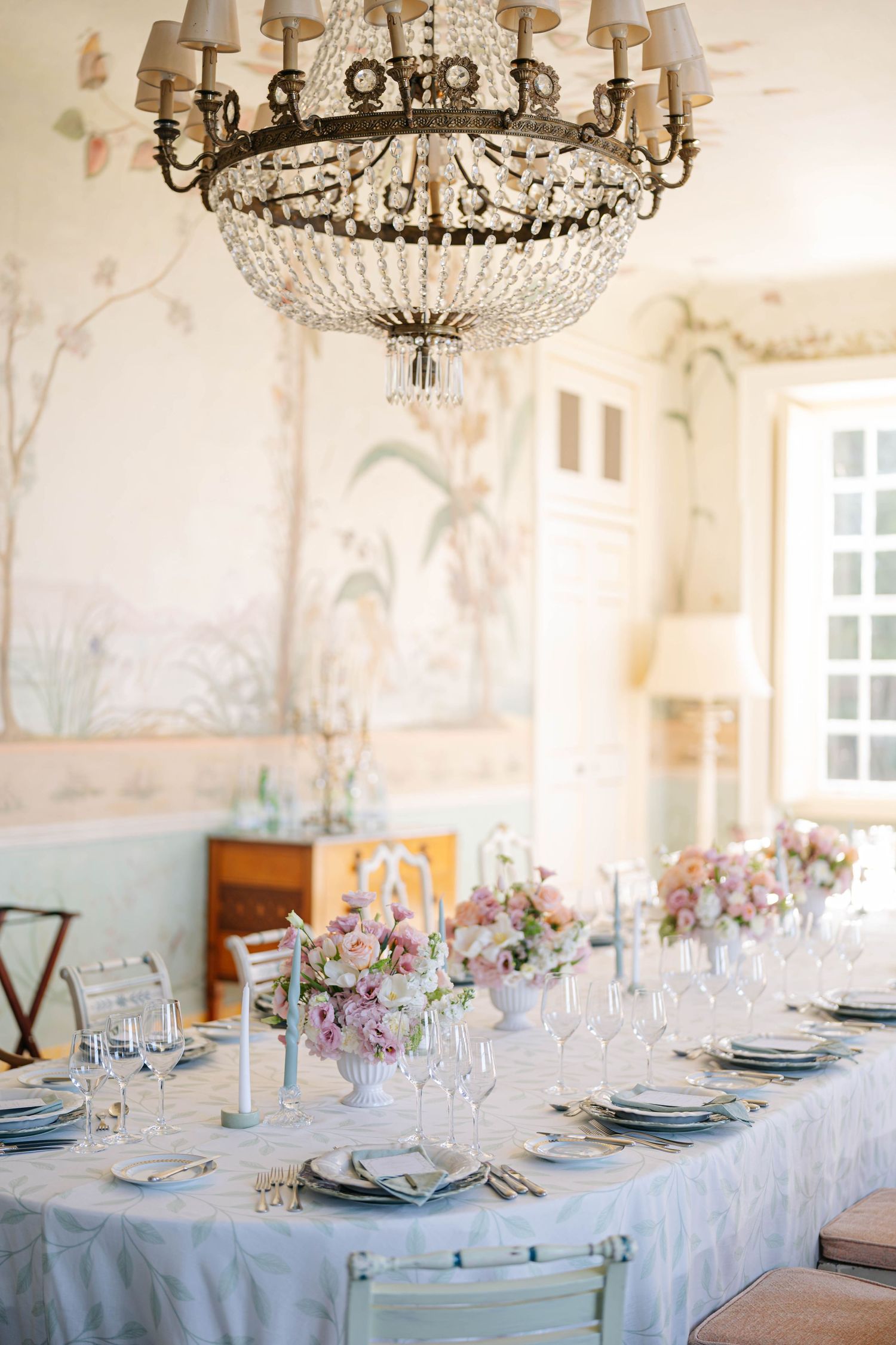 Luxurious banquet tables adorned with crystal chandeliers and blue linens in a room with hand-painted pastoral murals.