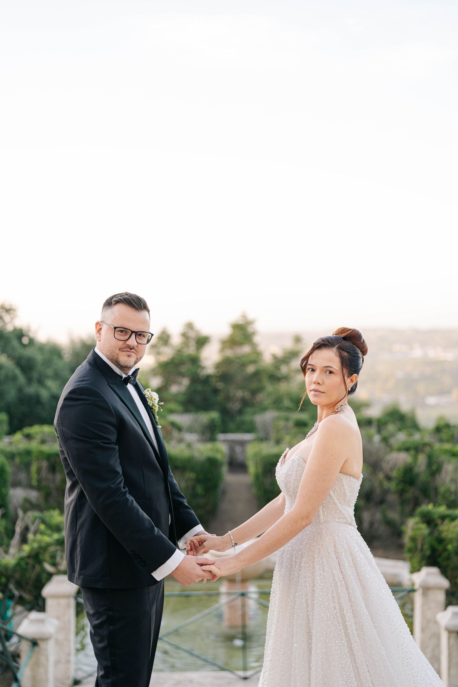 A couple in wedding attire holds hands on an outdoor terrace with lush greenery and scenic views in the background.
