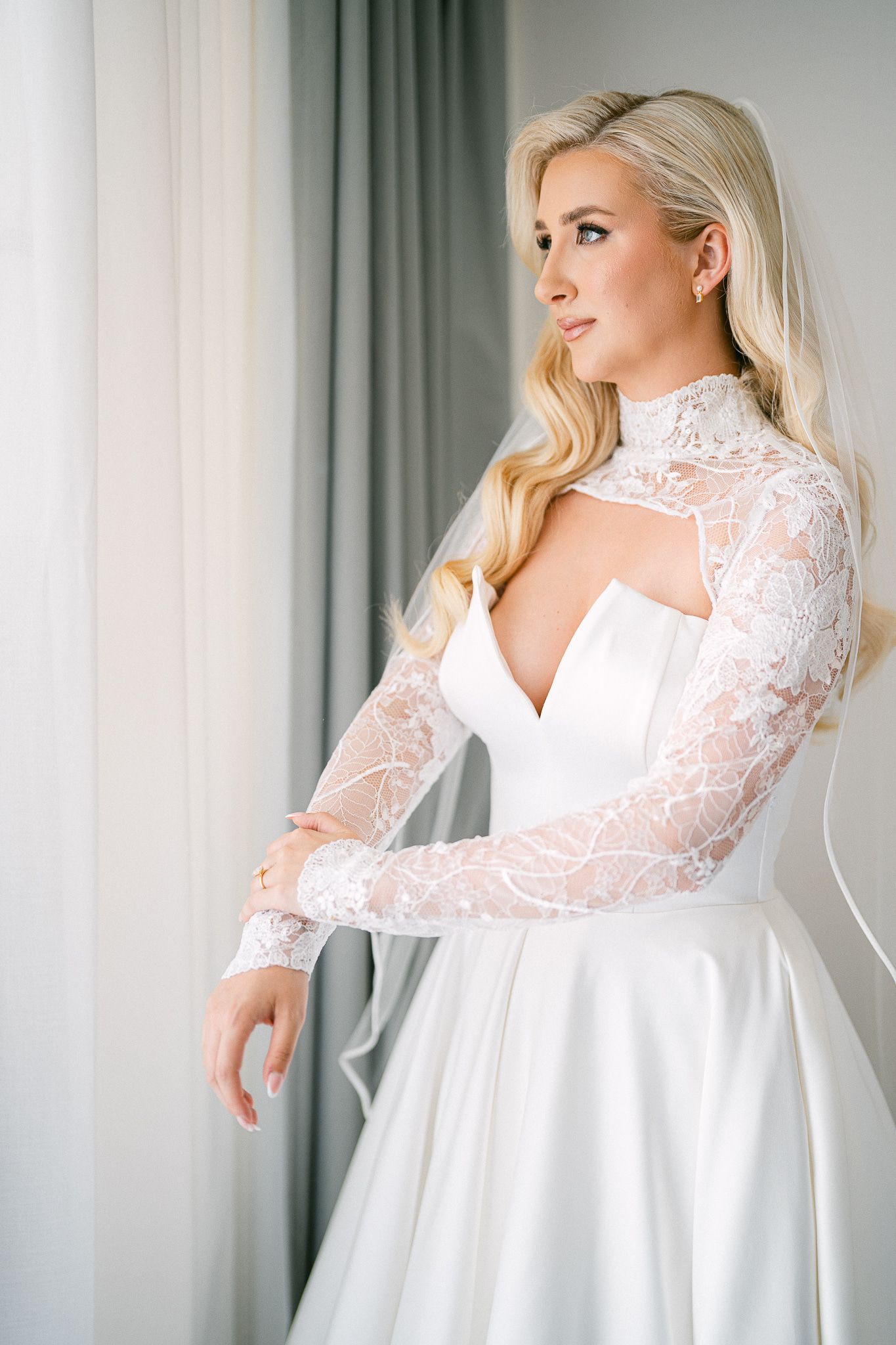 Side profile of bride in modern wedding dress with sheer lace sleeves against soft window light.