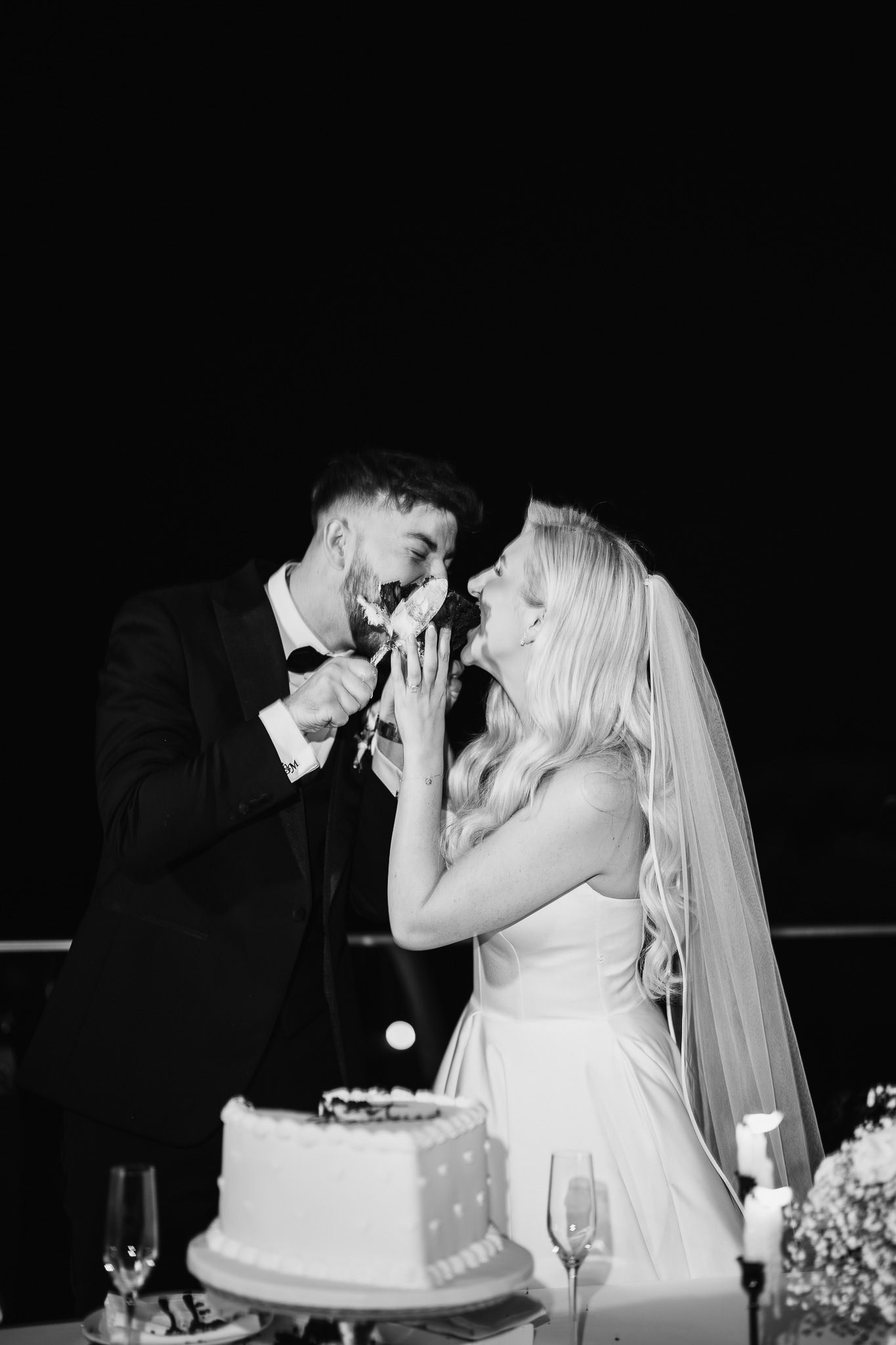 Black and white capture of couple feeding each other wedding cake with laughter.