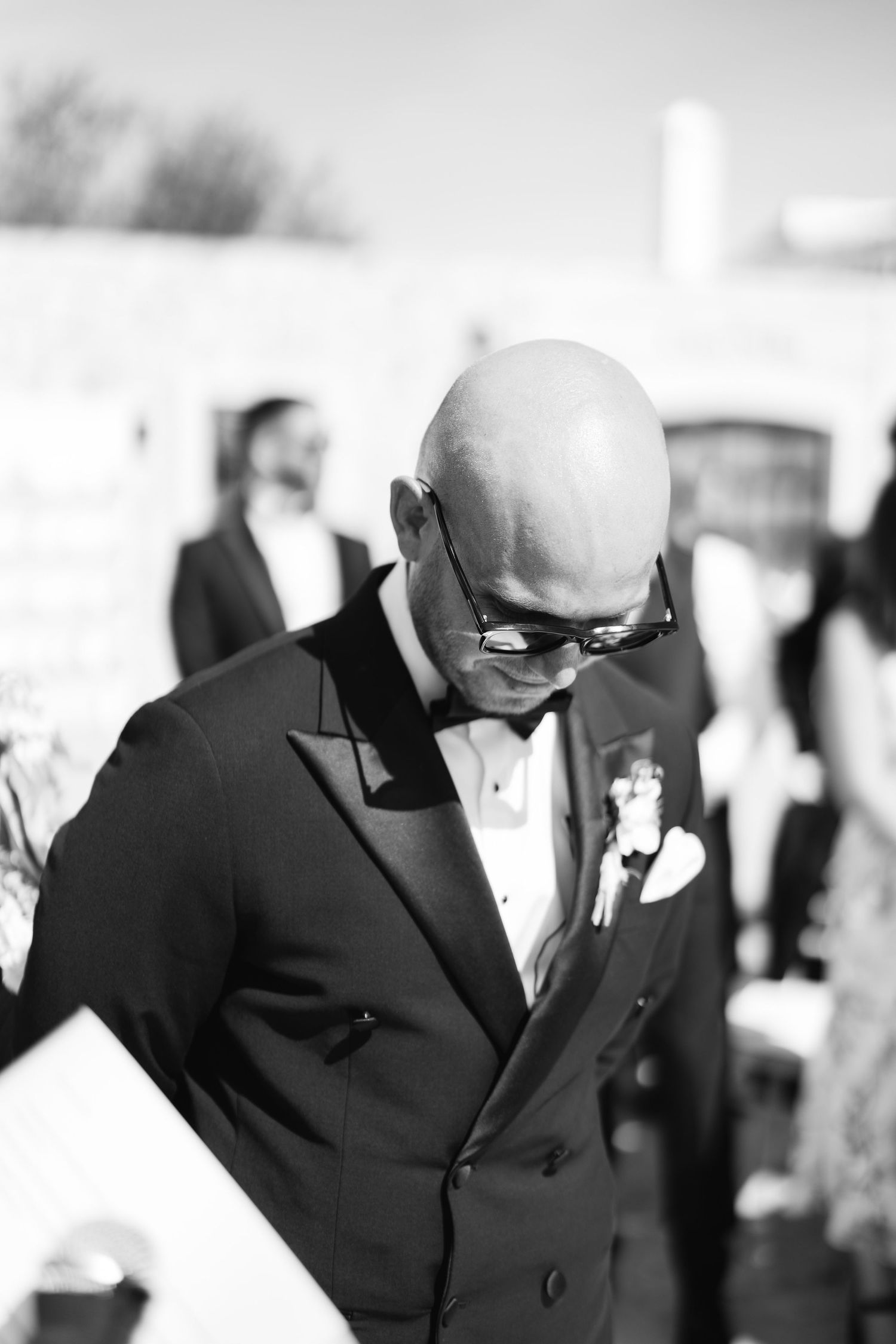 Wedding attendee in black tuxedo and bow tie at formal outdoor celebration.
