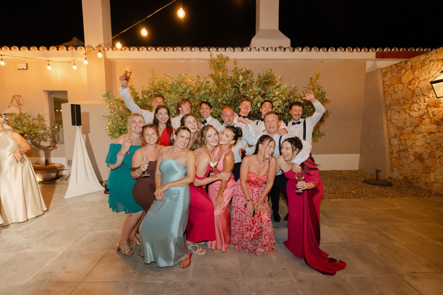 Wedding party guests in colorful dresses sit together at an outdoor reception under string lights.