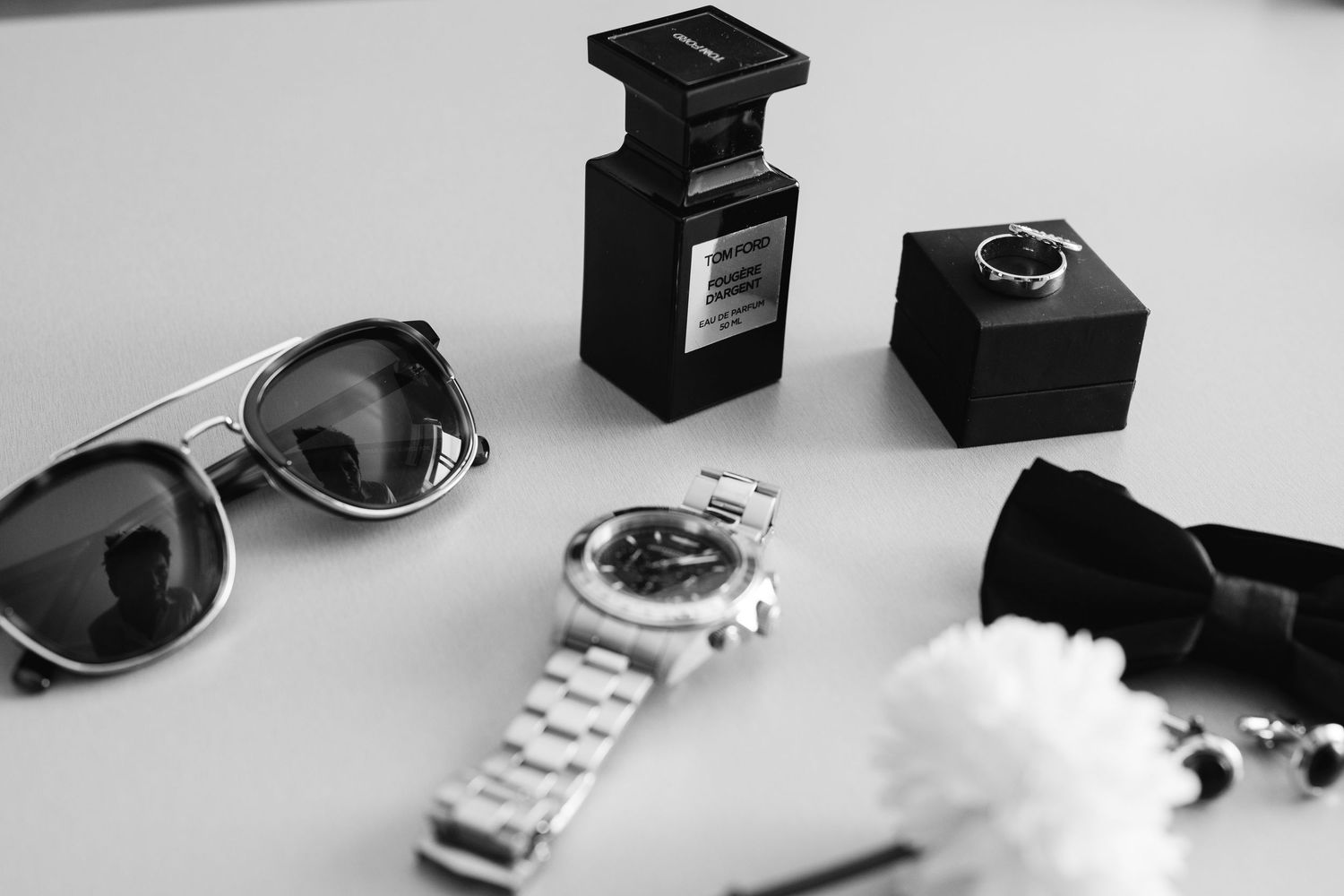 Luxury accessories including sunglasses, perfume bottle, watch and bow tie arranged on white surface.