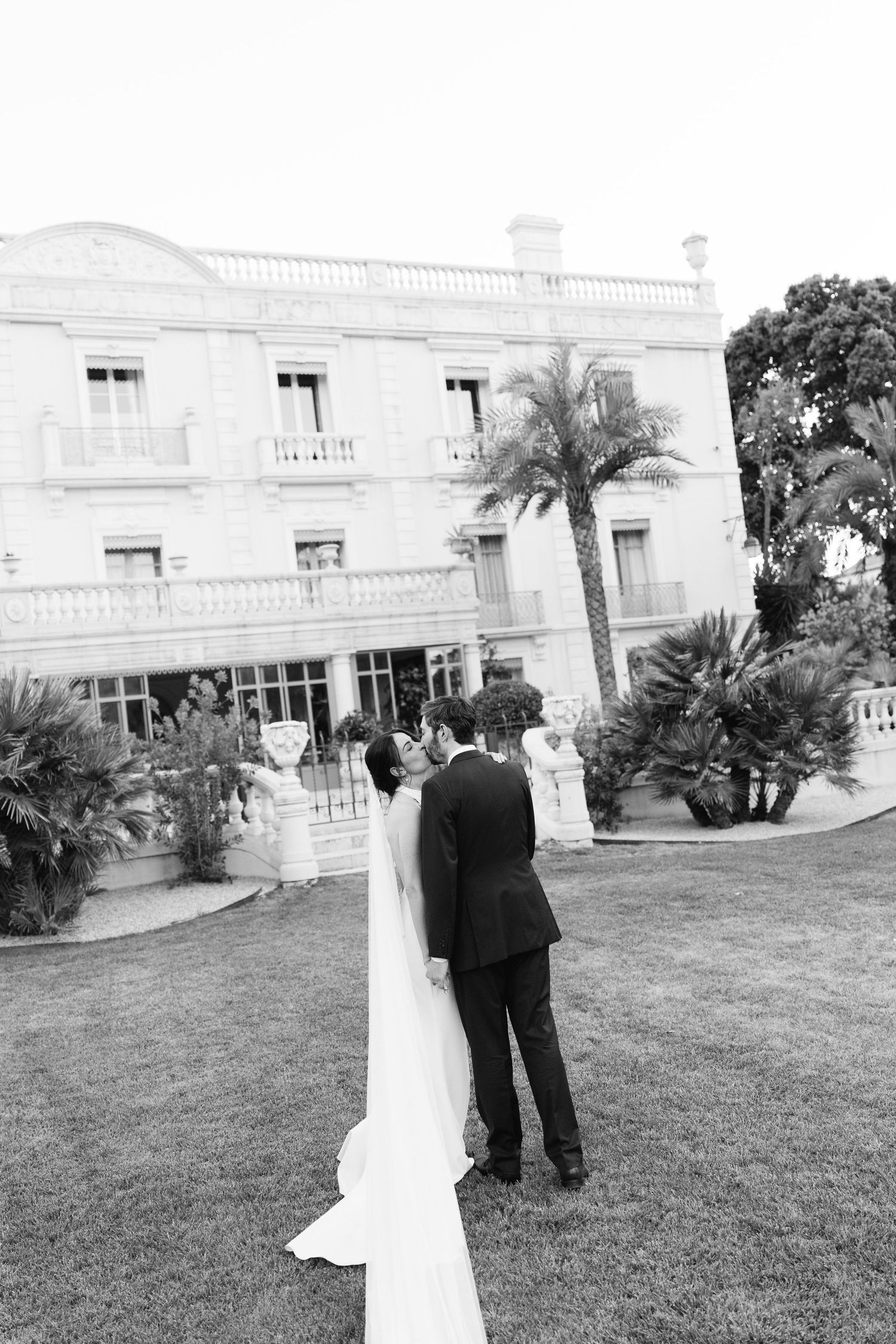Black and white photo of bride and groom standing on lawn before elegant mansion with palm trees.