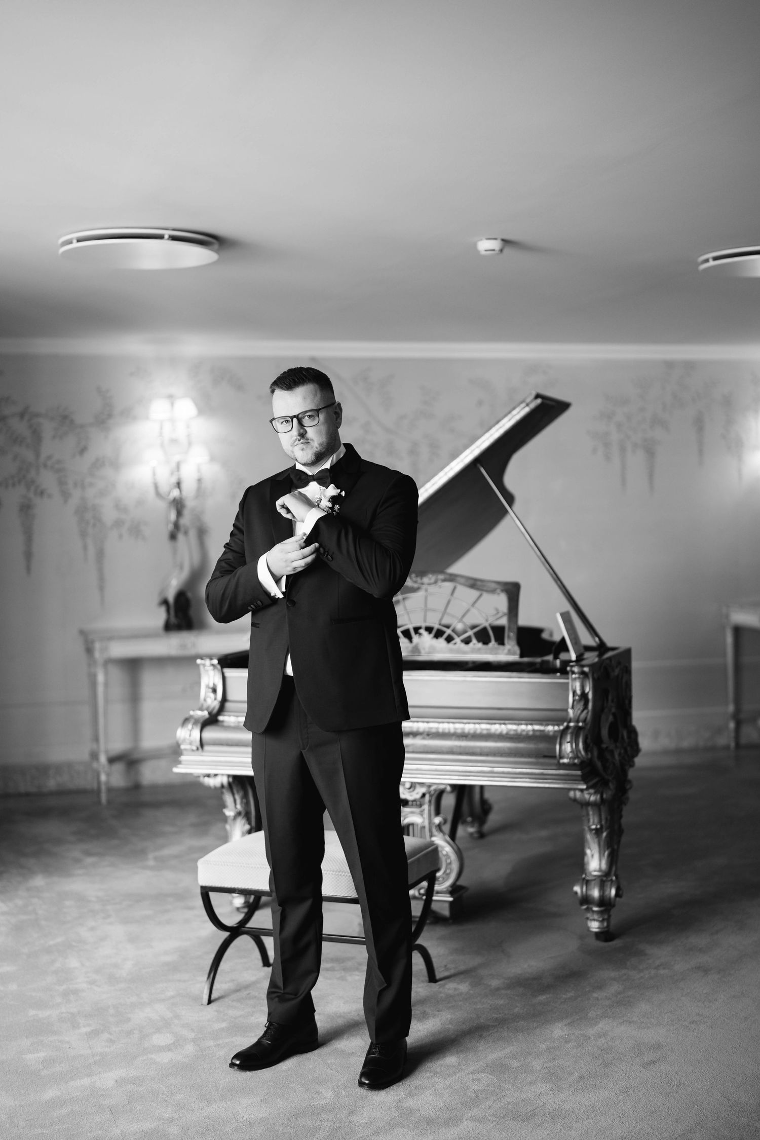 Black and white photo of a person in a suit standing in front of a grand piano in an elegant room.