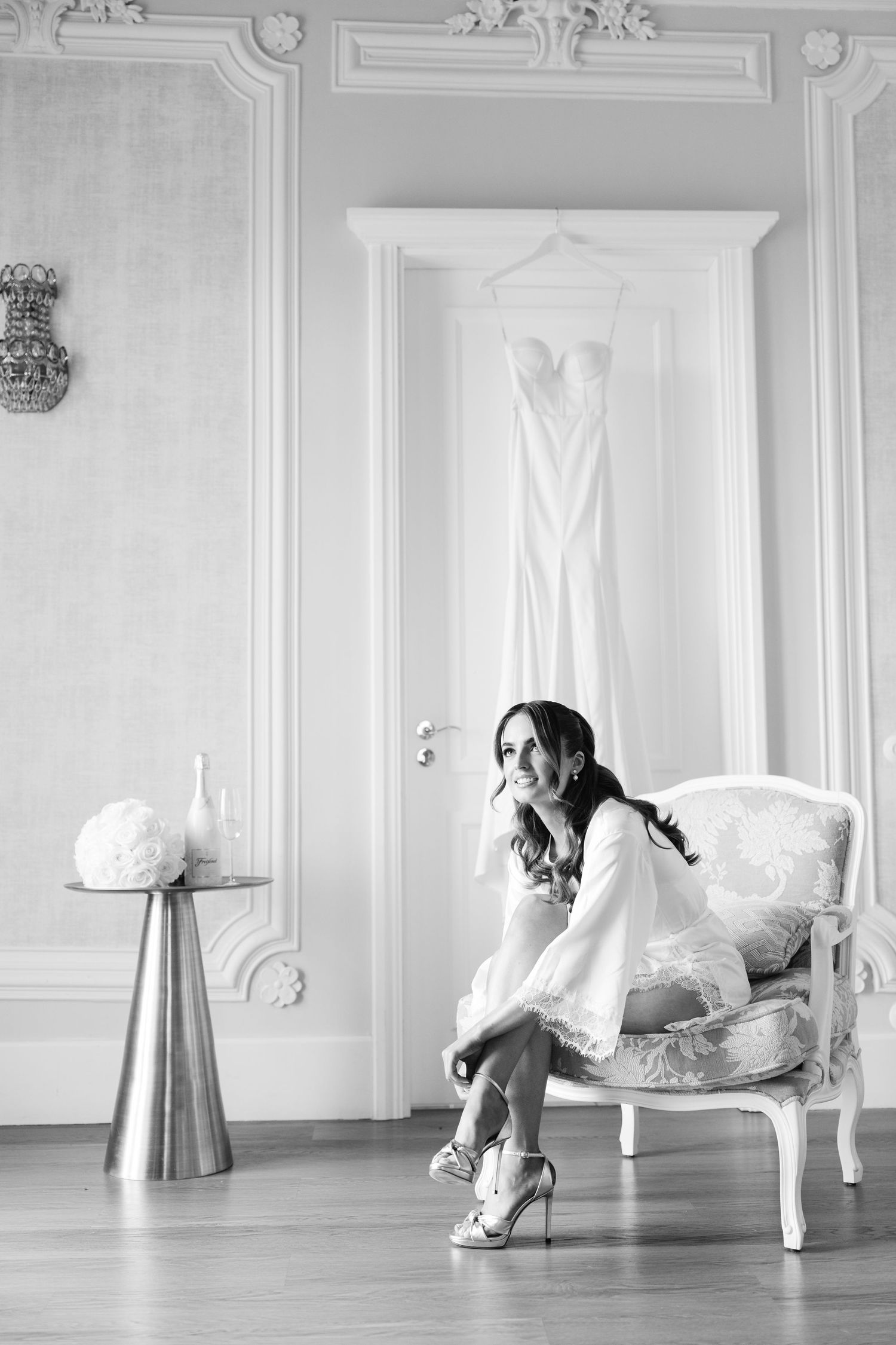 Black and white photo of woman sitting in vintage chair with wedding dress hanging in background.