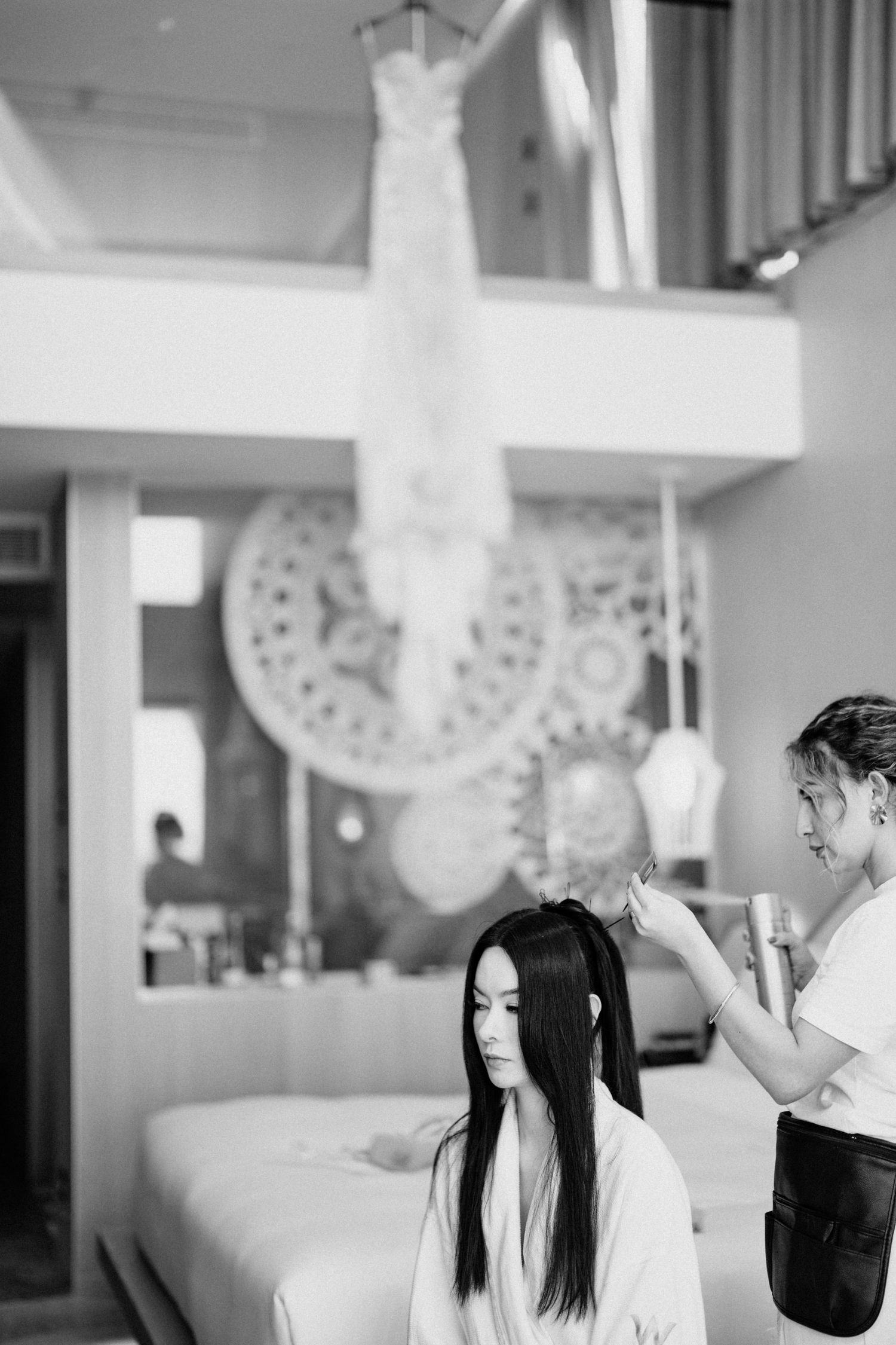 Stylist working on bride's hair preparation with wedding dress hanging in background.