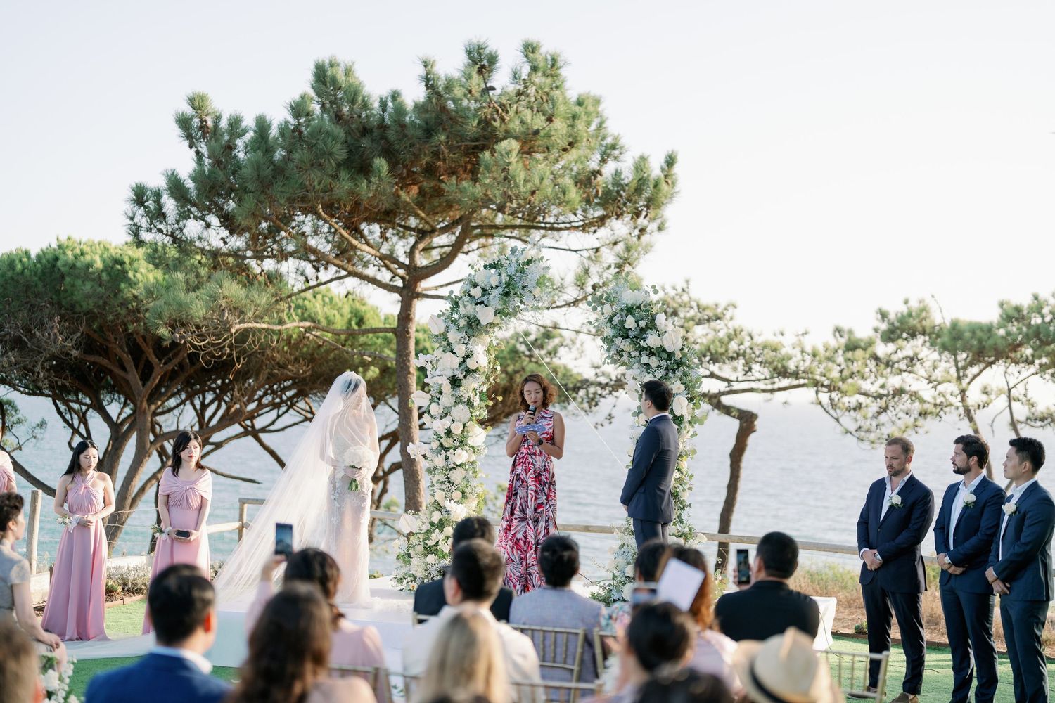 Outdoor wedding ceremony under pine trees with guests watching the proceedings.